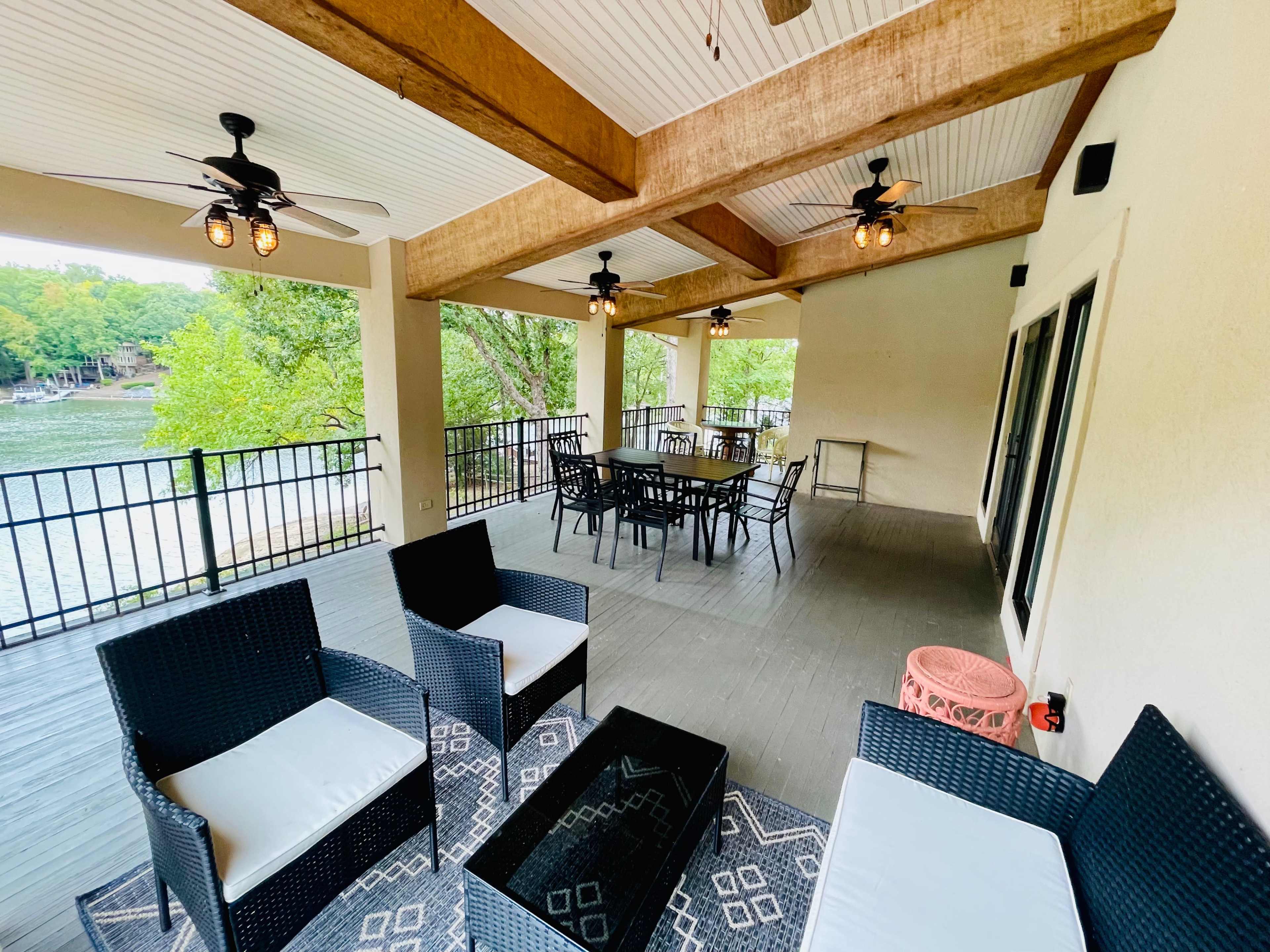 A covered outdoor patio features black wicker furniture, a dining table, and several ceiling fans, overlooking a body of water and surrounded by greenery.