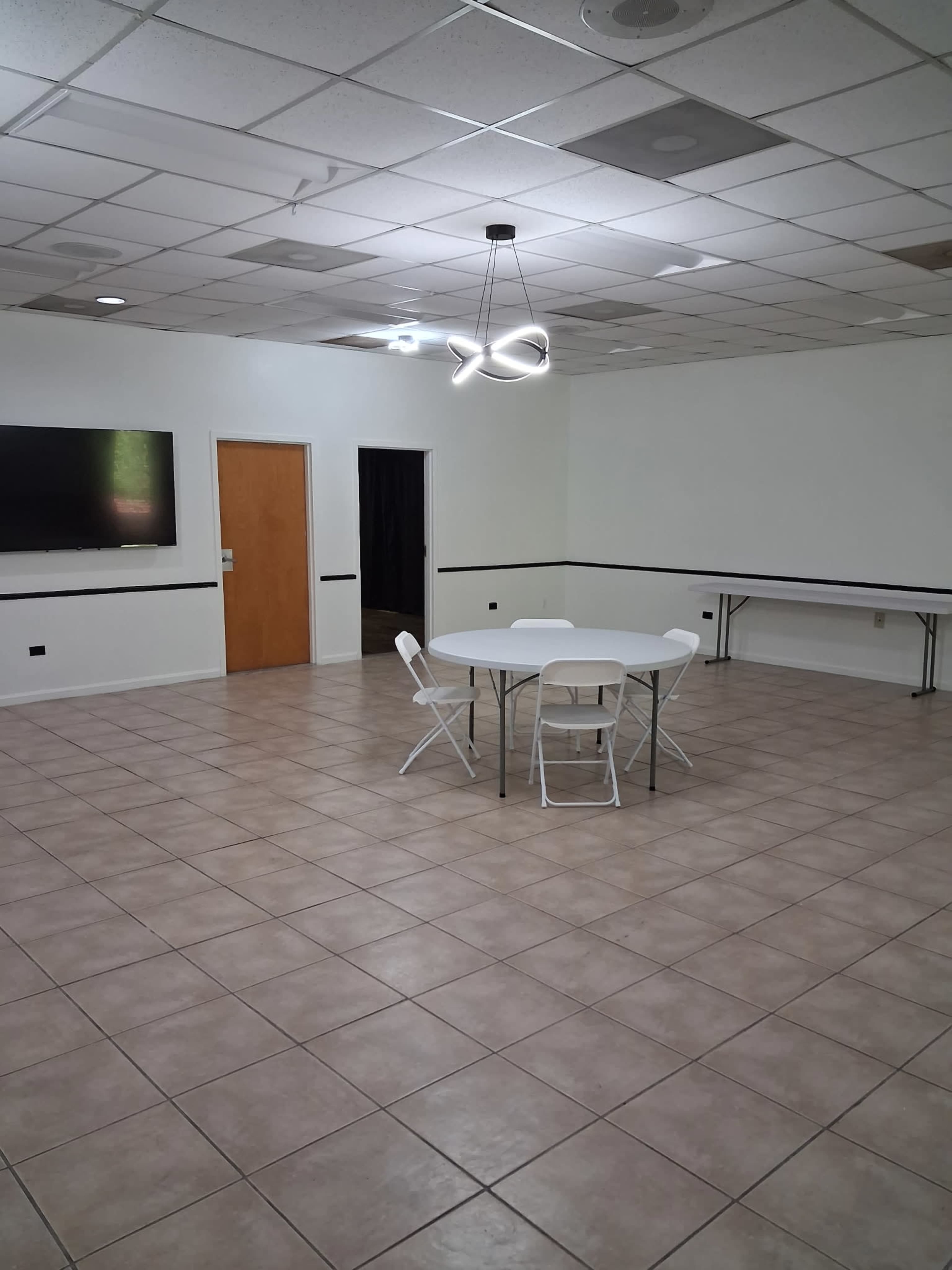 The image shows a spacious room with tan tile flooring, featuring a round table surrounded by four white chairs, a wall-mounted screen, and a door leading to another area.