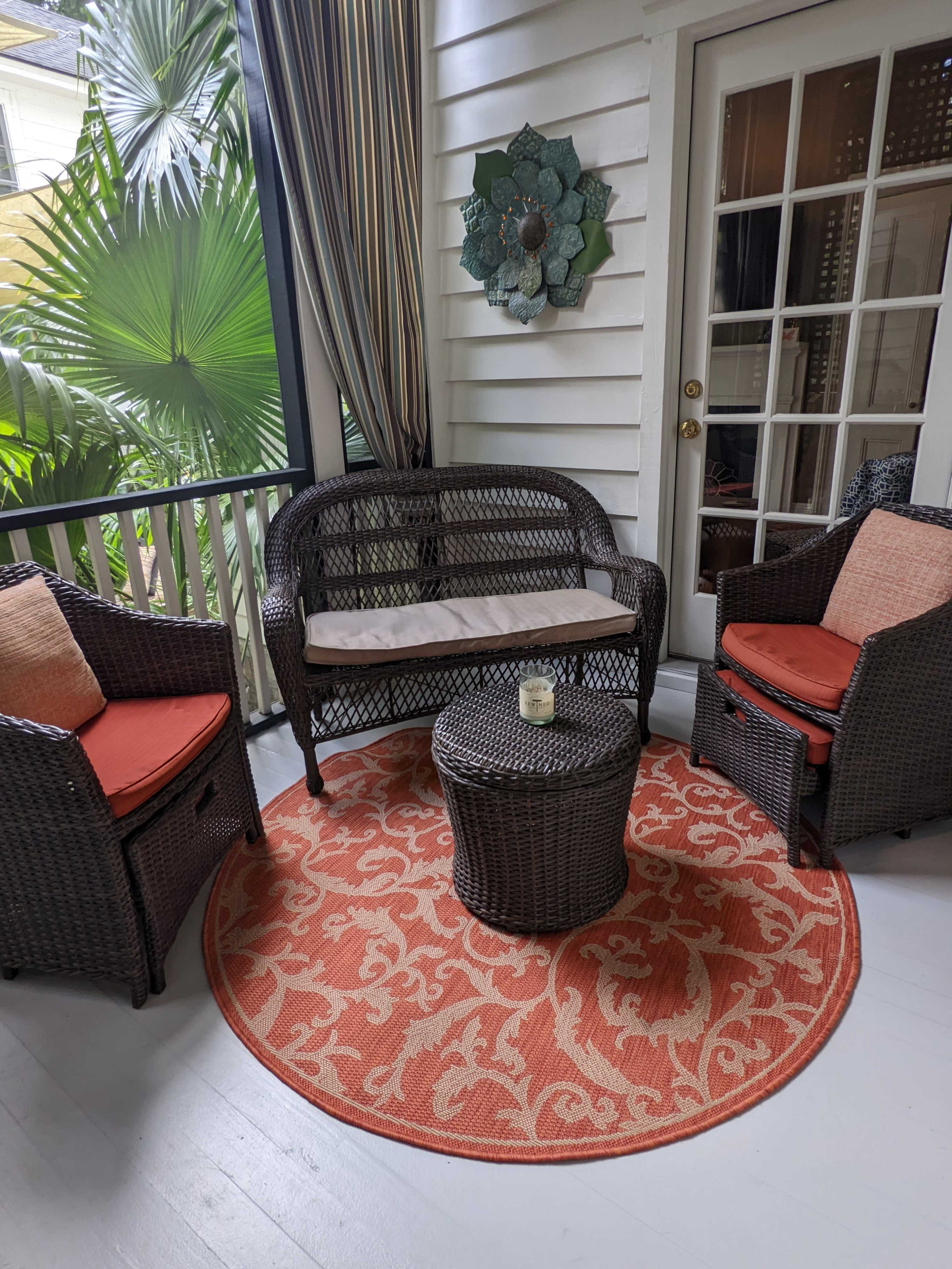A small sitting area is set up on a porch with two wicker chairs and a round table, surrounded by plants and decorated with an orange patterned rug.
