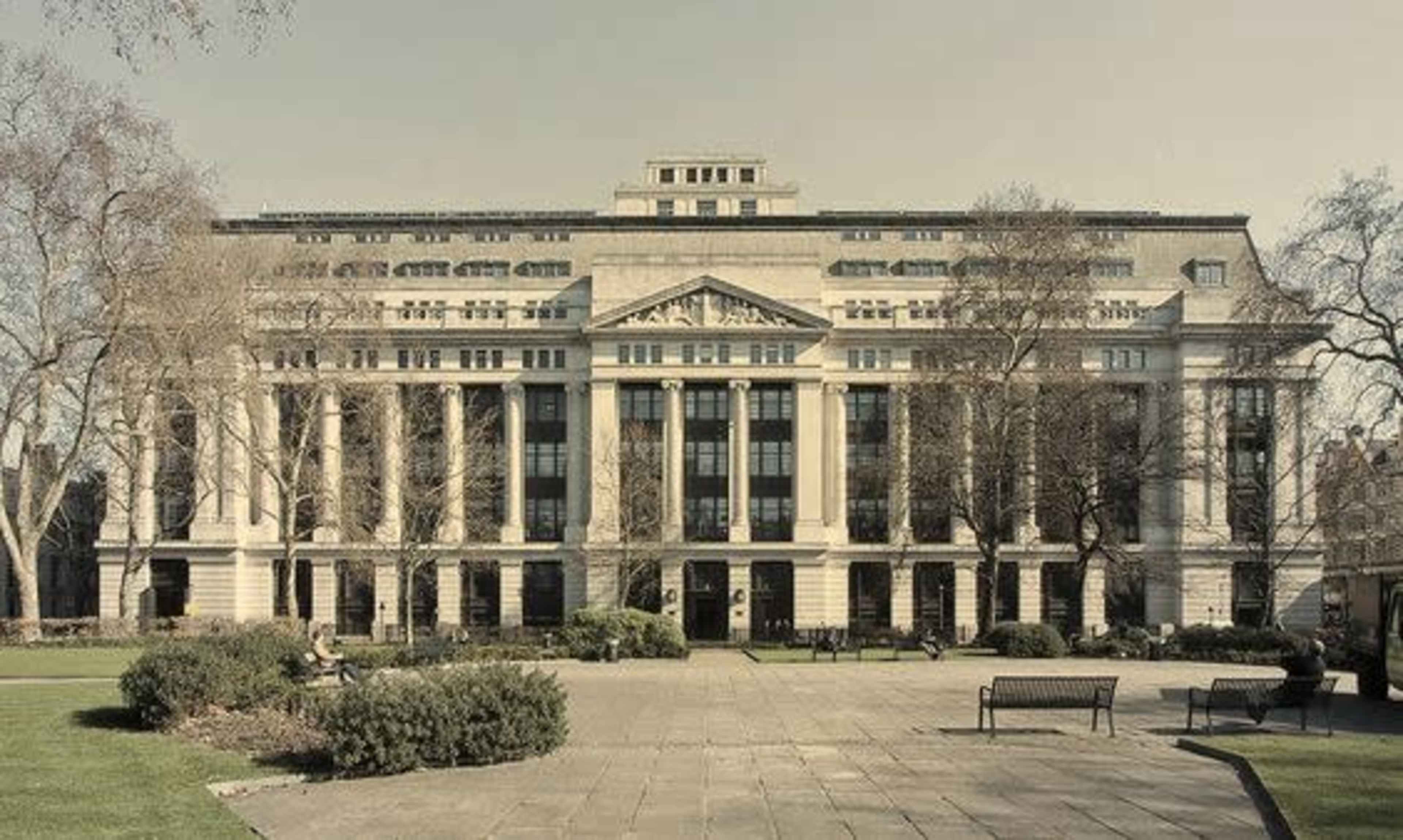 A large, classical-style building with columns and a symmetrical facade, surrounded by a park area with benches and trees.