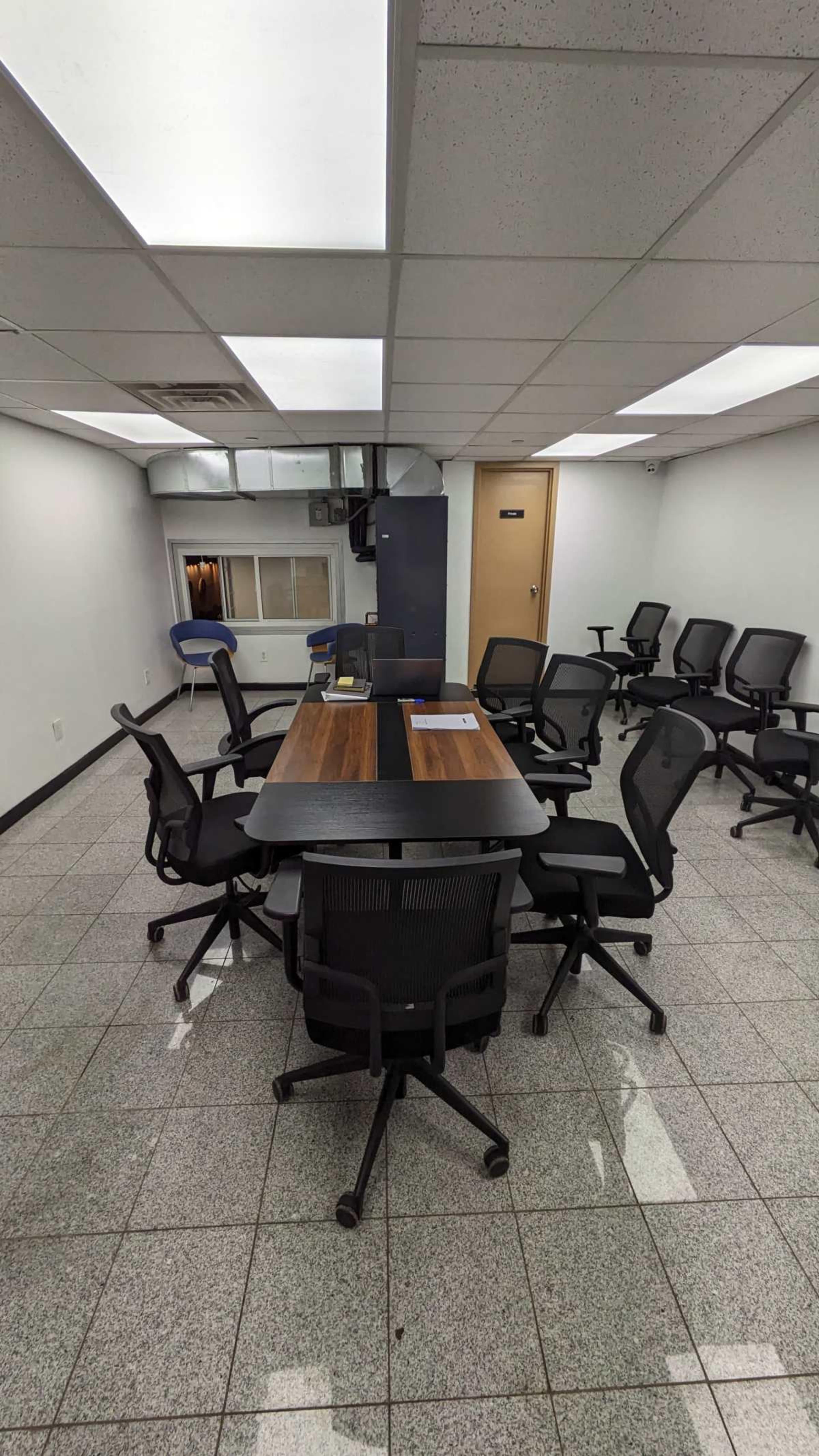 A conference room features a central wooden table surrounded by black office chairs, with additional empty chairs lining the walls.