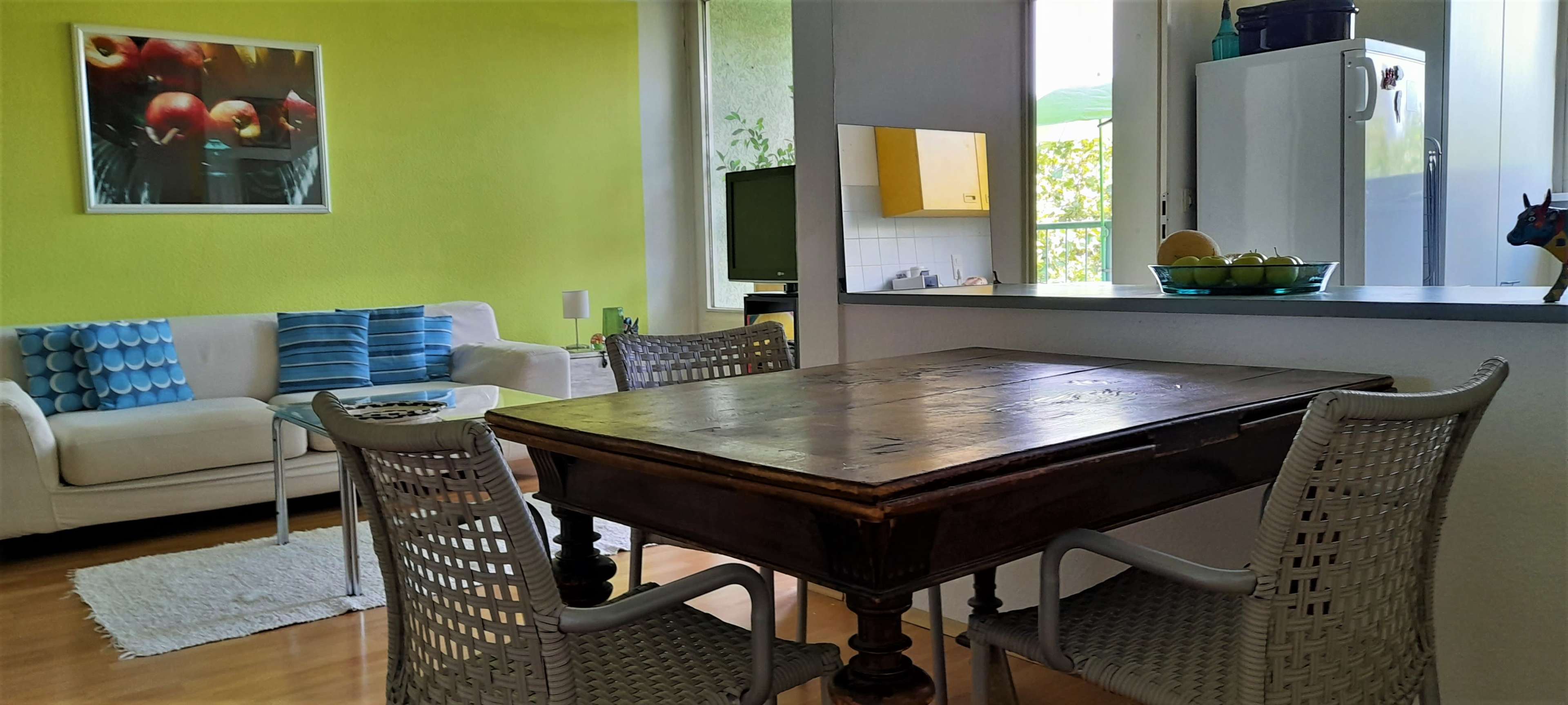 The image depicts a cozy dining area with a wooden table surrounded by four chairs, adjacent to a living space featuring a sofa and colorful decor.
