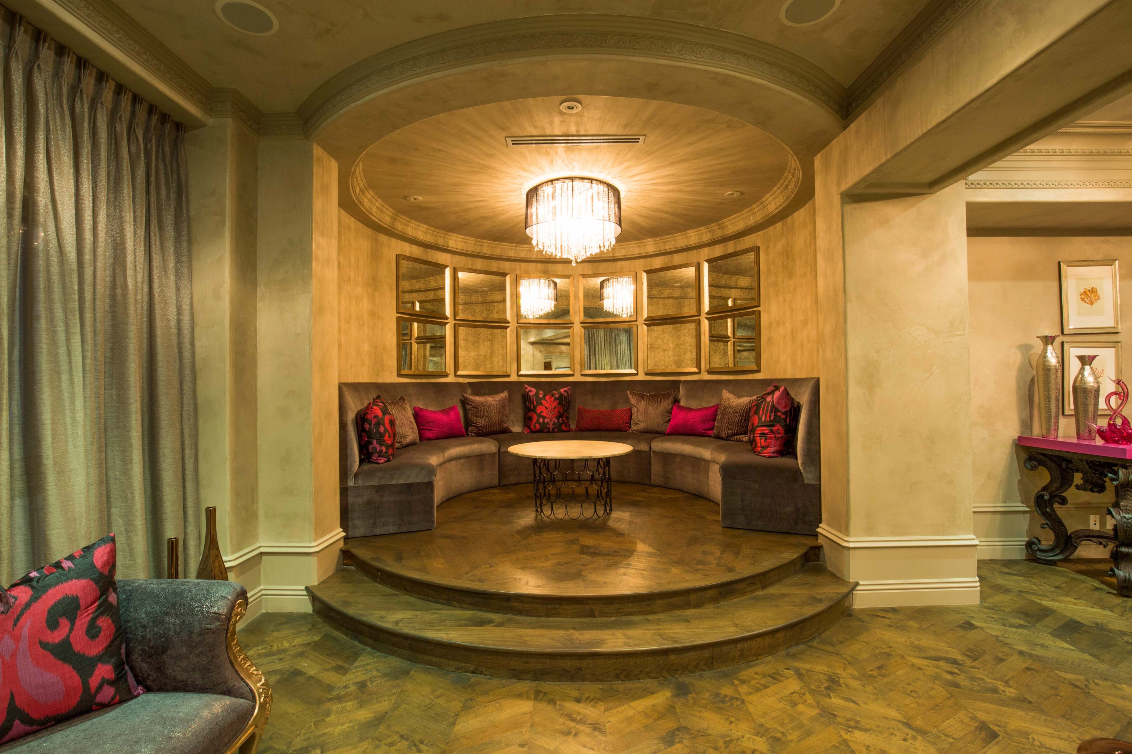 A circular seating area with a round table, surrounded by plush cushions and adorned with decorative mirrors, all illuminated by a chandelier above.