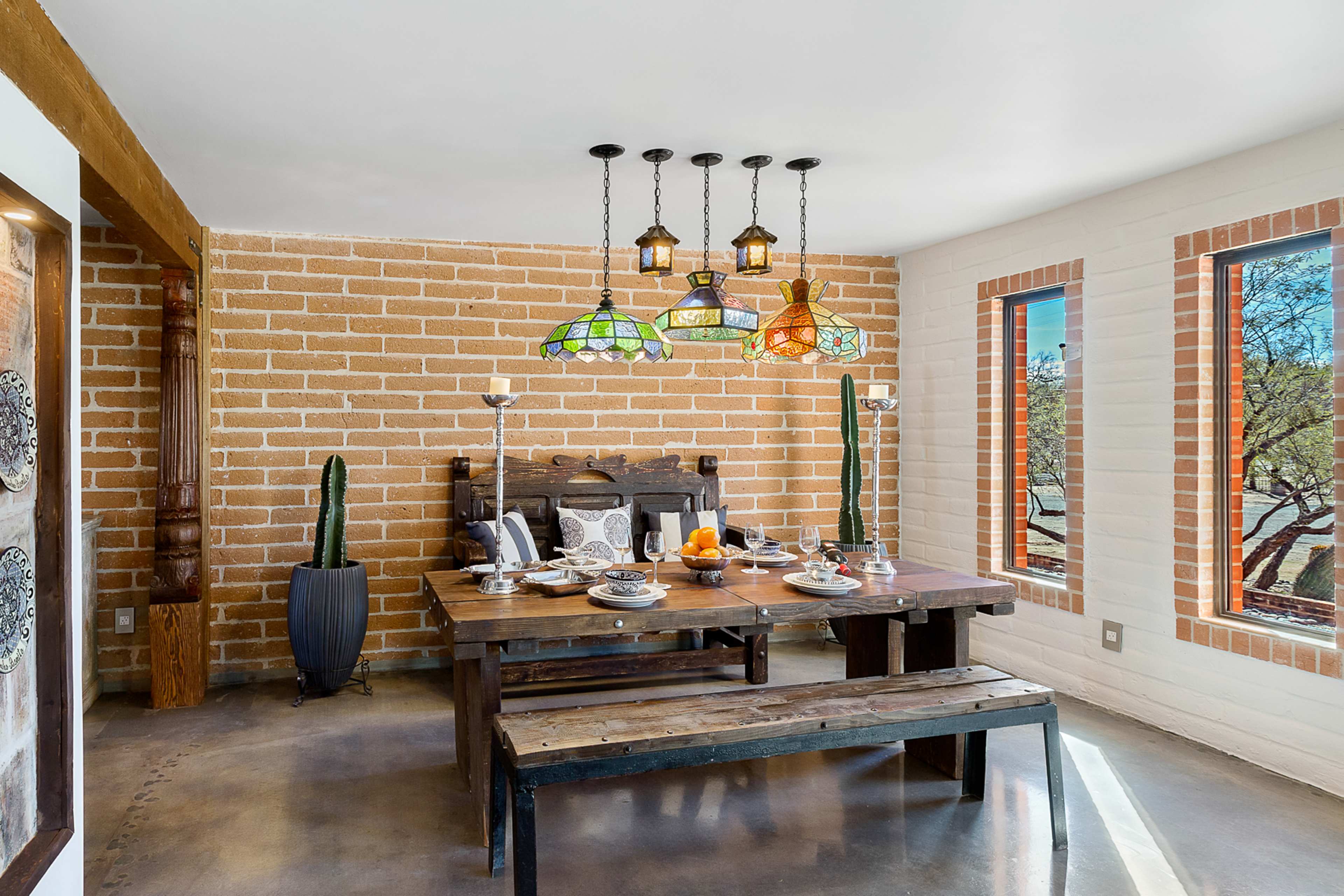 A dining area features a wooden table set with plates, surrounded by colorful pendant lights and large windows.