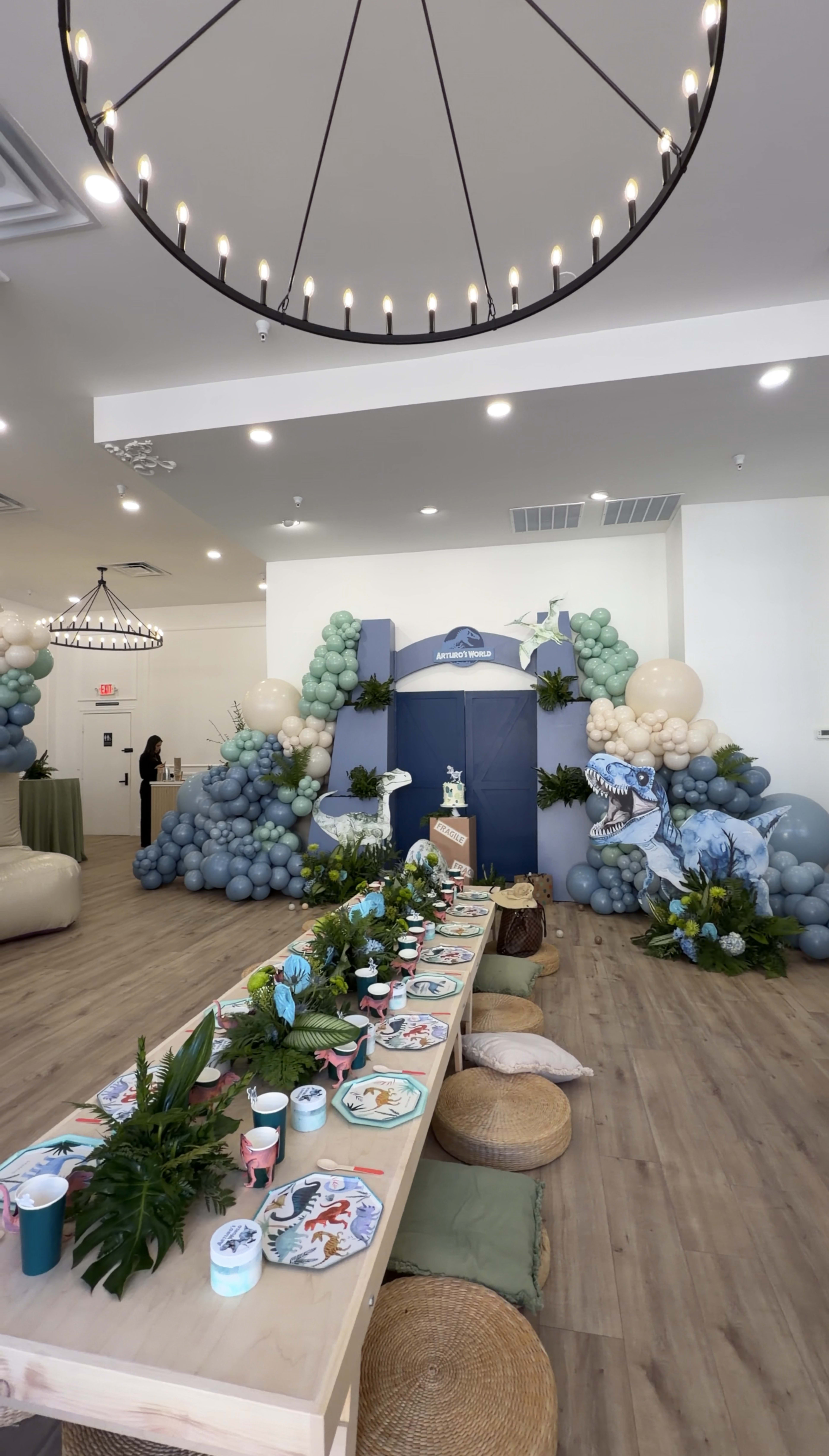 The image shows a decorated event space with a long table set up for a celebration, surrounded by balloon arrangements in various colors and themes, and featuring a backdrop with a blue mural.