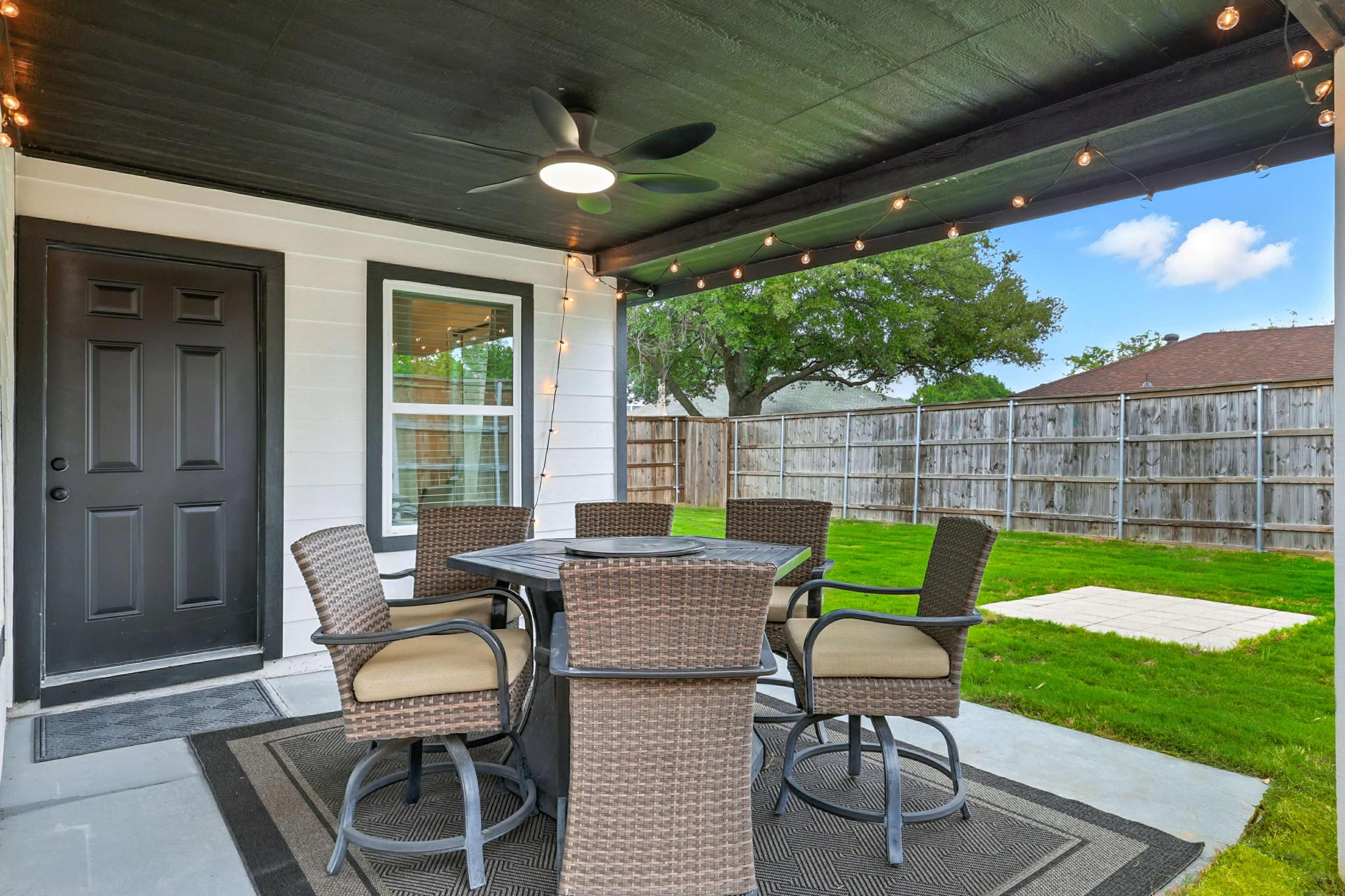 The image shows a covered outdoor patio with a dining table and chairs, decorative string lights, and a grassy yard enclosed by a wooden fence.