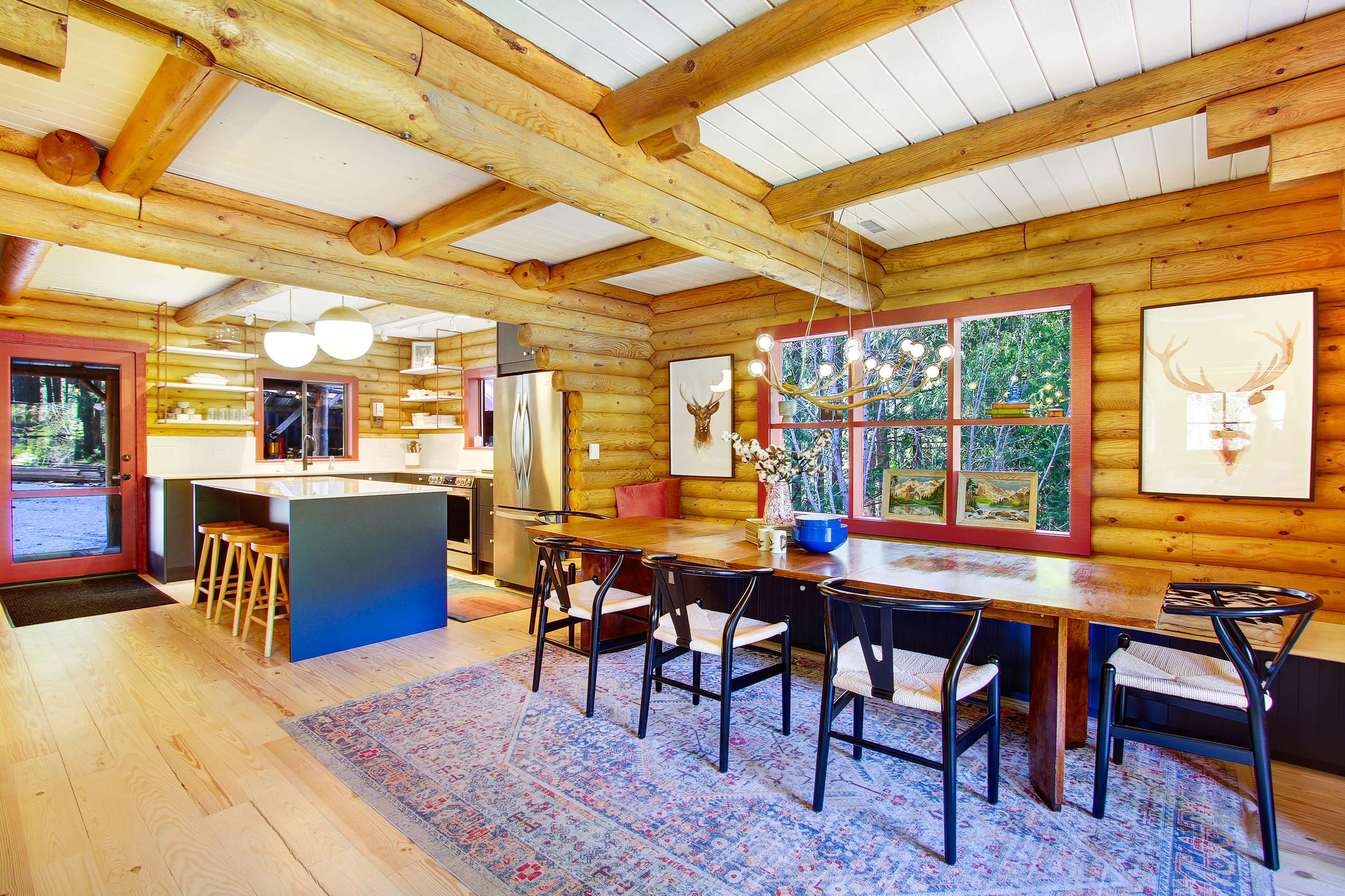 The image shows a log cabin kitchen and dining area with a long wooden table, modern appliances, and large windows facing a natural view.