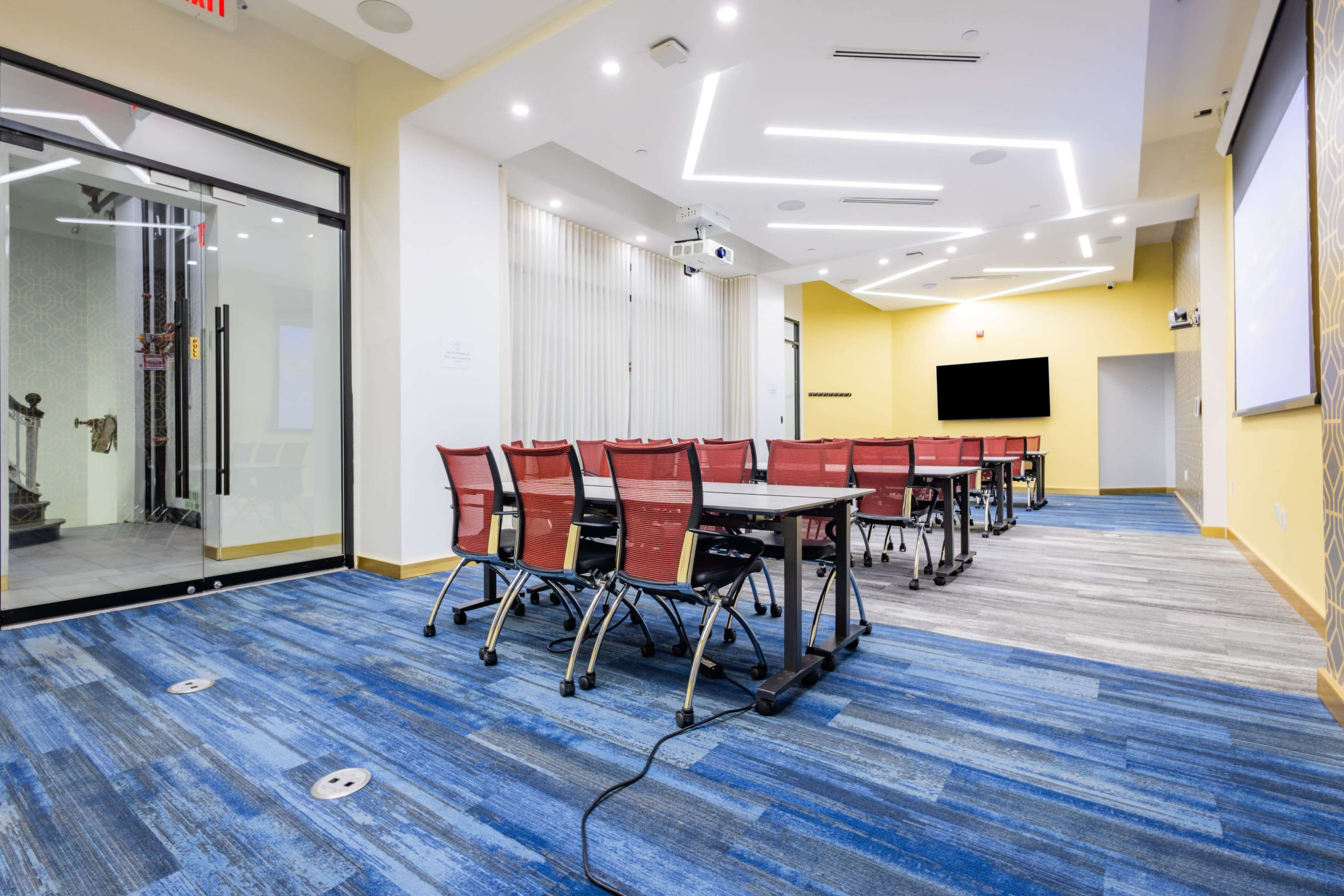The image shows a modern conference room with a long table, chairs, a projector, and a large screen on the wall.