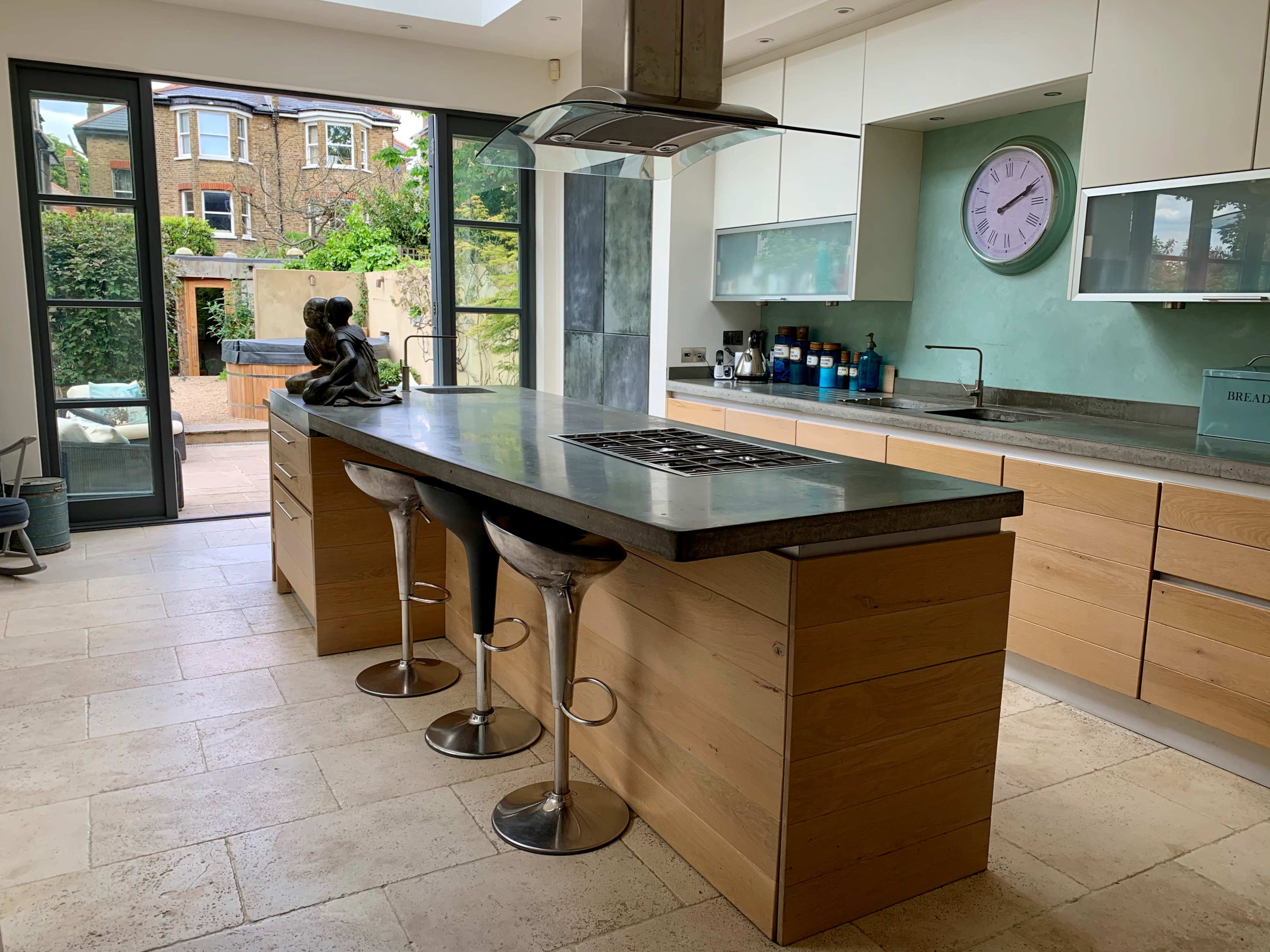 The kitchen features a central island with a stovetop, modern stools, and large windows overlooking an outdoor space.