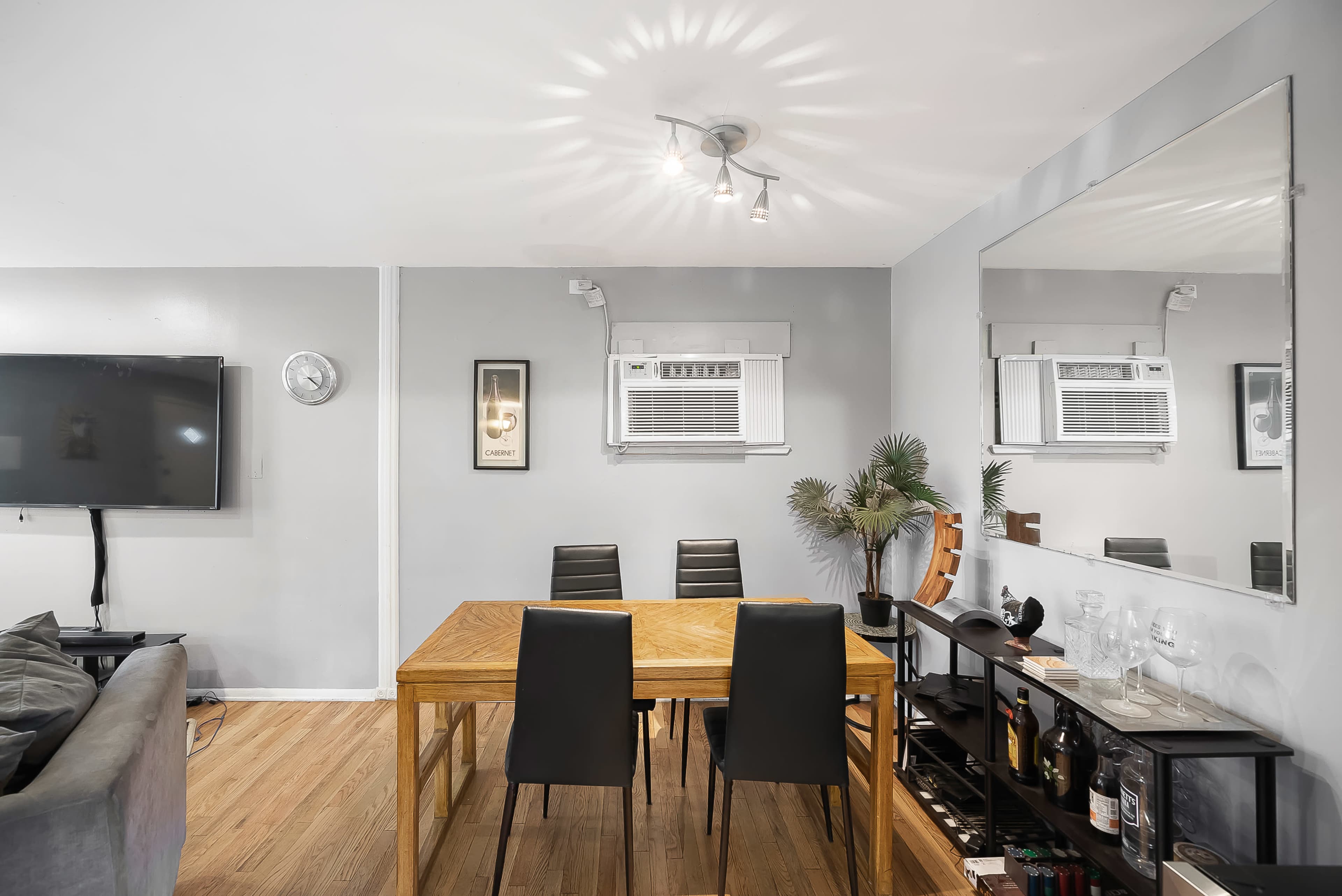 The image shows a dining area with a wooden table, four black chairs, a wall-mounted television, and a mirror reflecting the space.