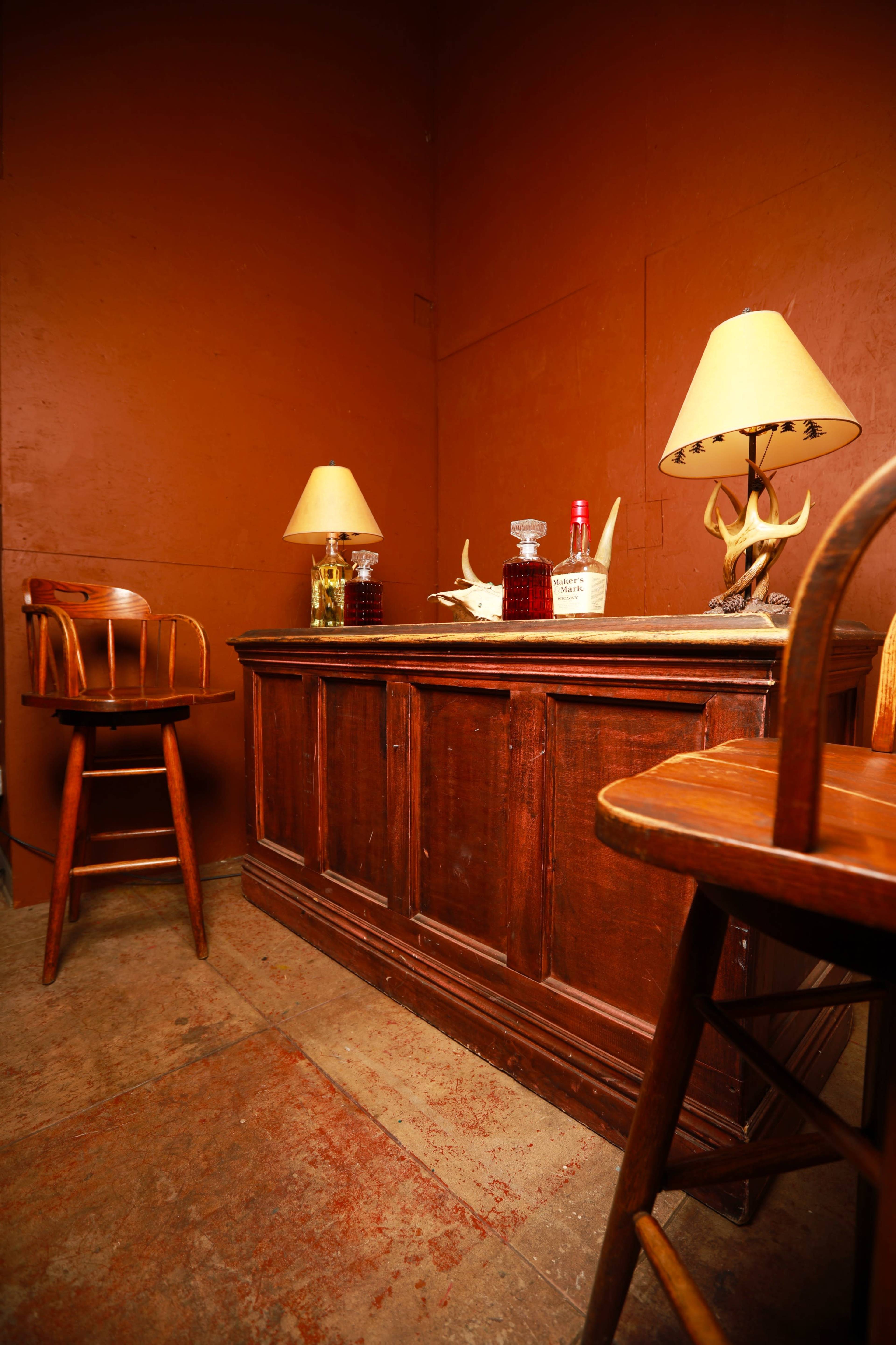 A wooden bar is positioned against an orange wall, flanked by two lamps and a pair of wooden stools.