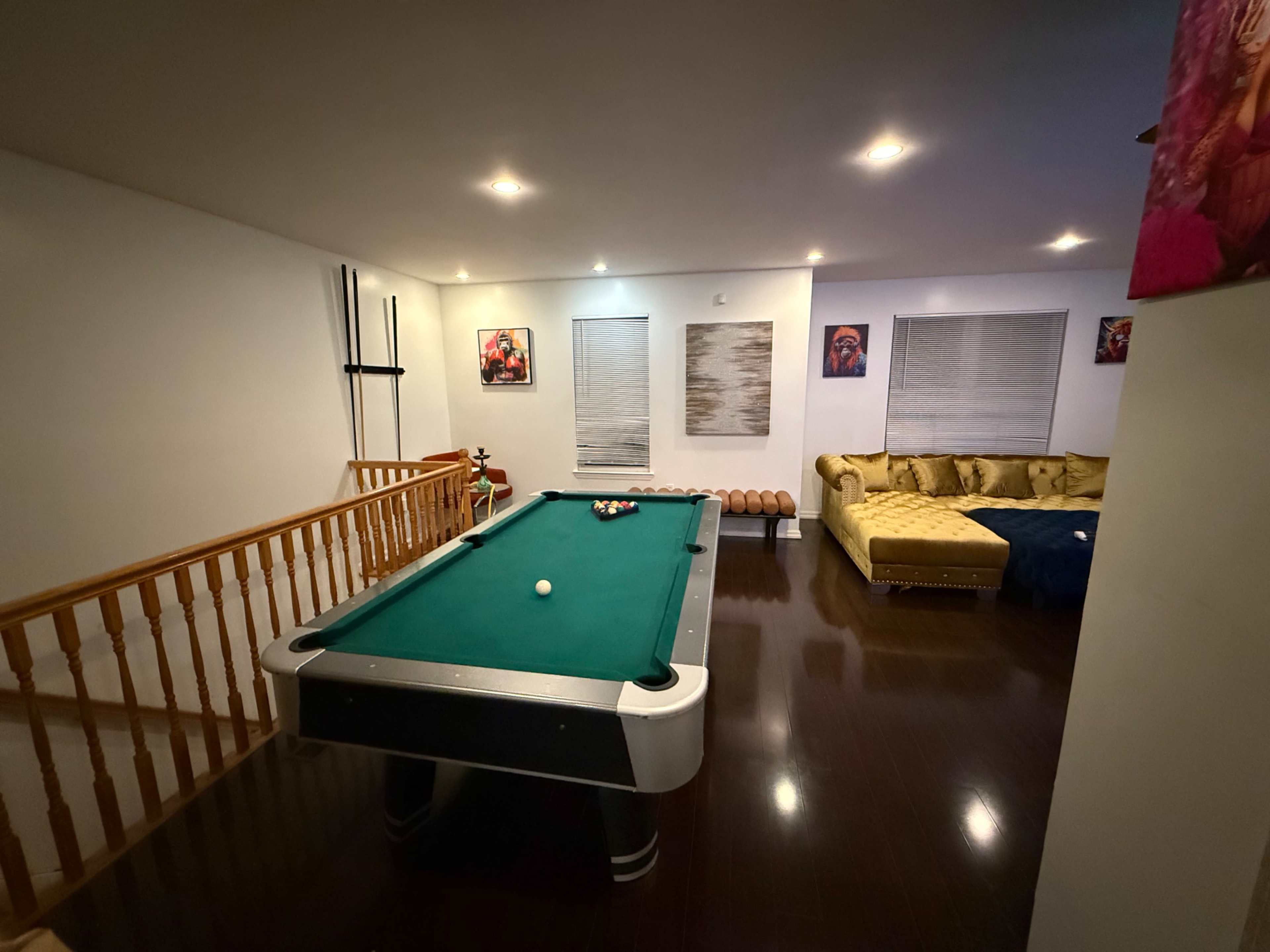 The image shows a game room with a green pool table, a yellow sectional sofa, and several framed artworks on the walls.
