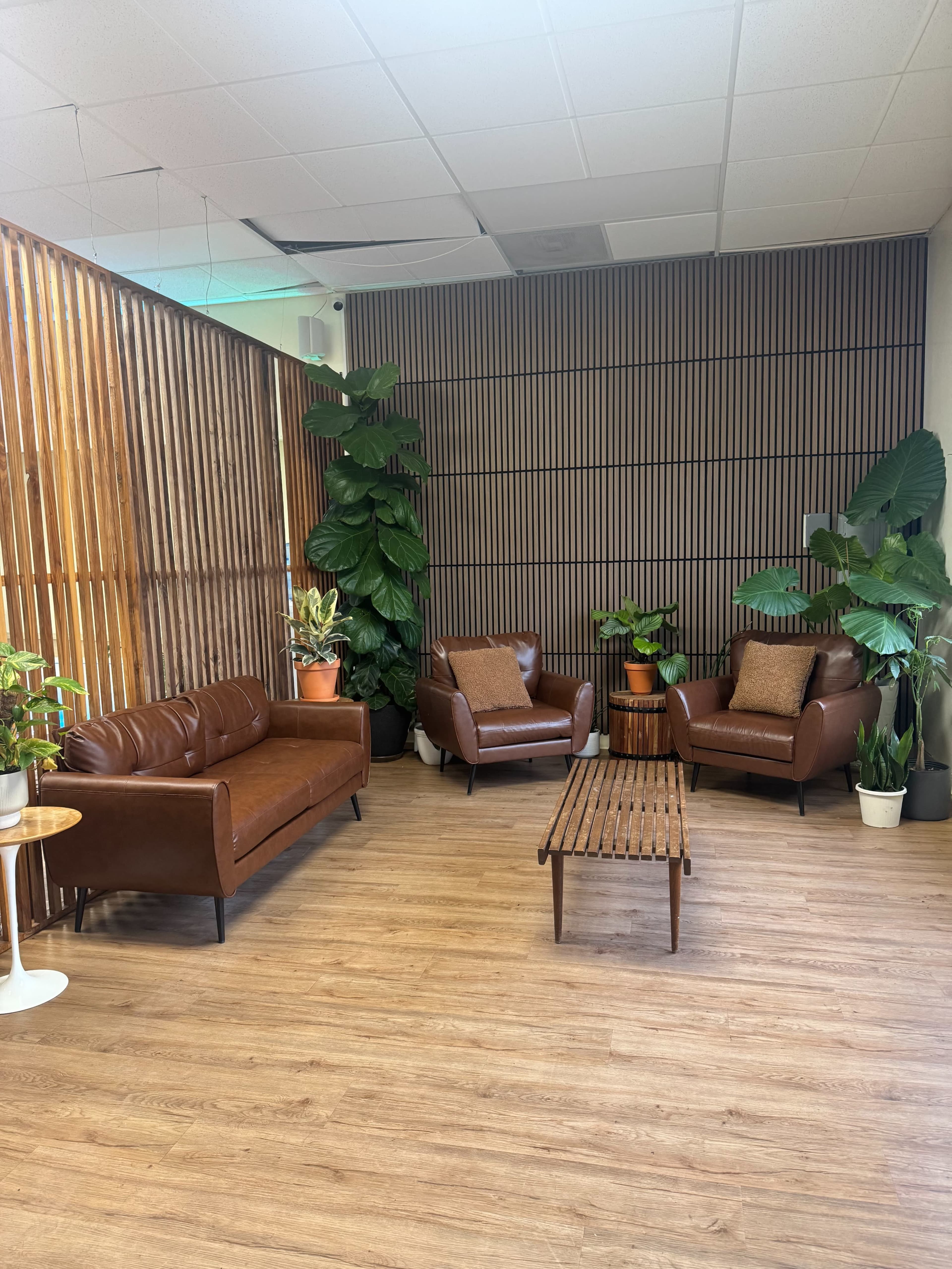 The image shows a cozy seating area with brown leather sofas, a wooden coffee table, and several potted plants against a wooden slatted wall.