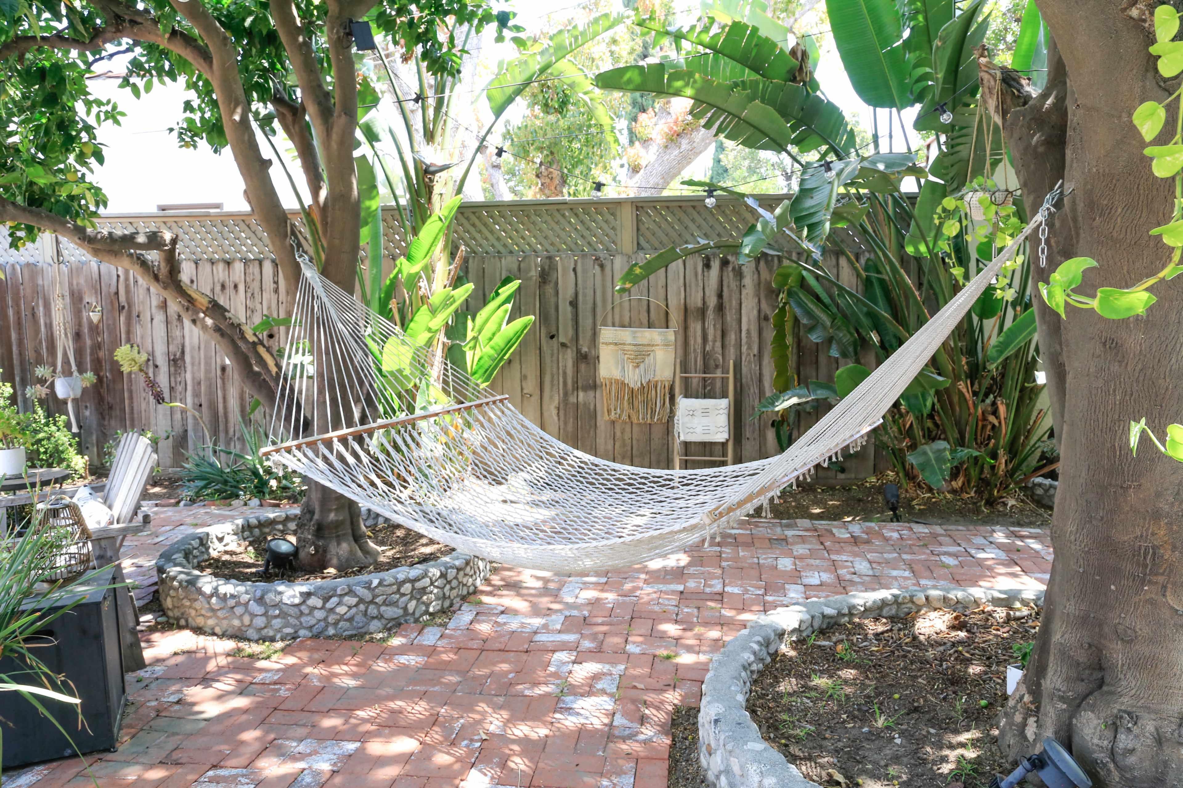 A hammock is suspended between two trees in a landscaped backyard surrounded by greenery and brick pathways.