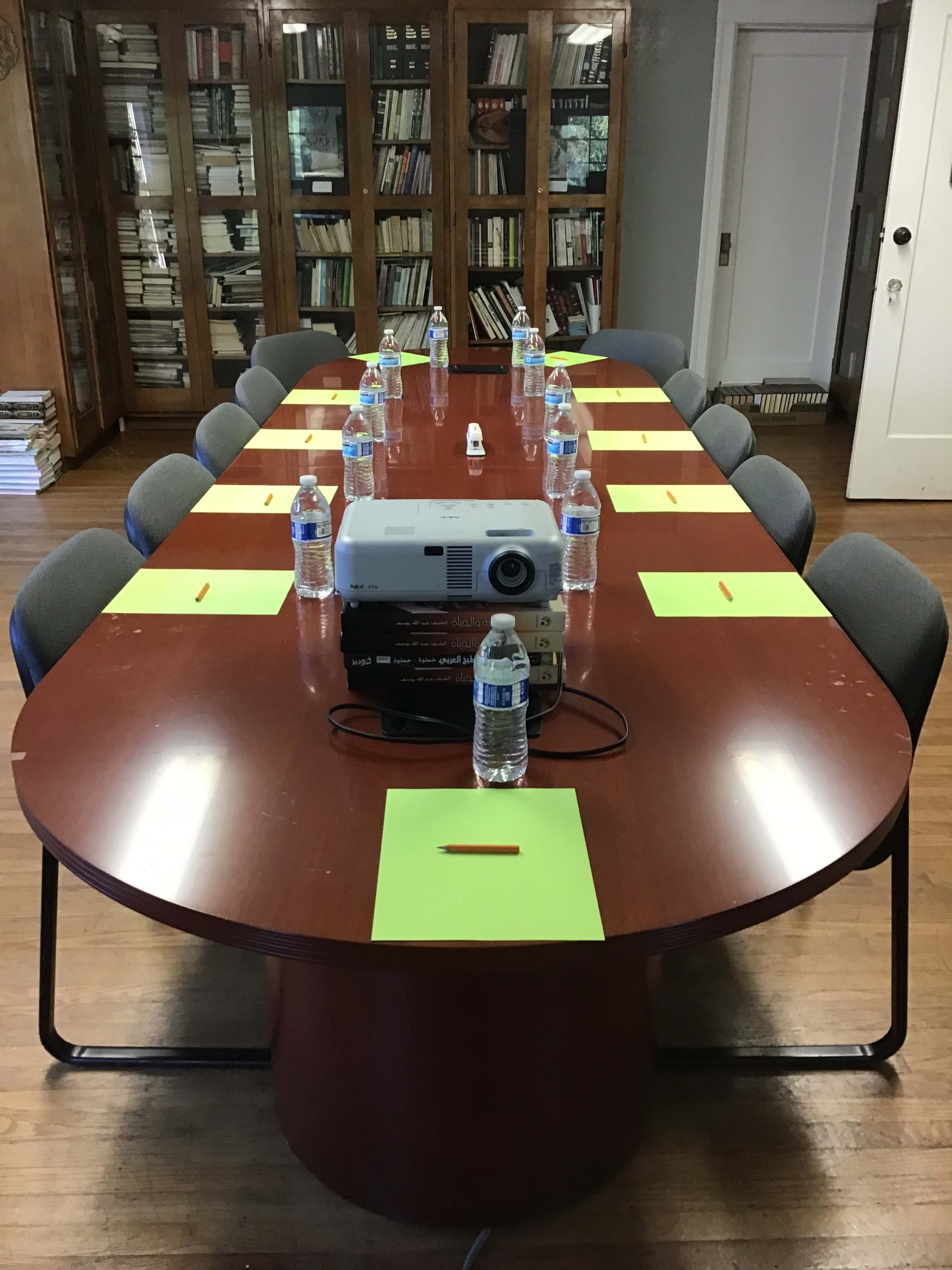 A large oval wooden conference table is set with water bottles and green paper place cards, with a projector positioned in the center.
