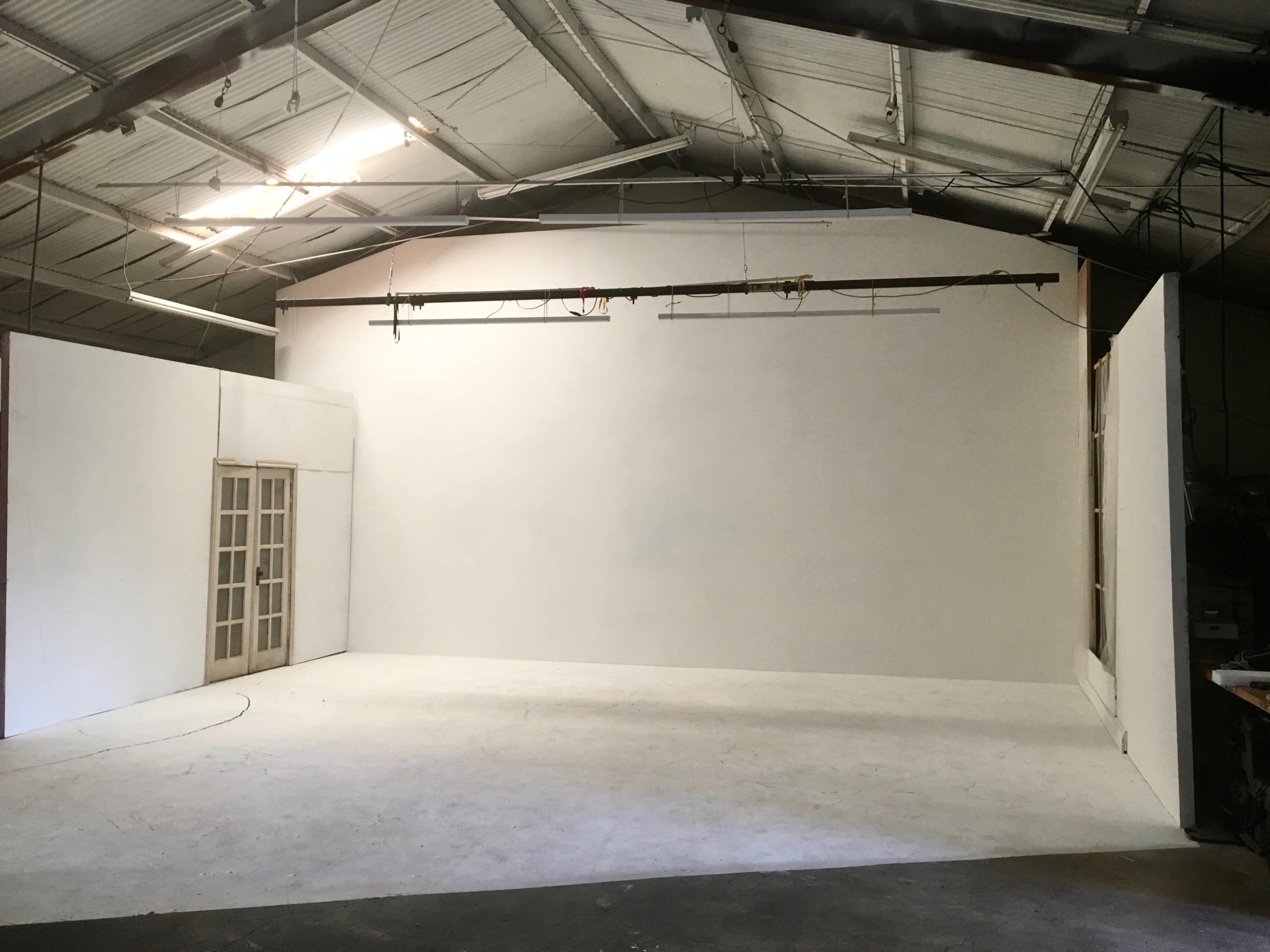 The image shows a large, empty studio space with white walls and a set of French doors on one side.