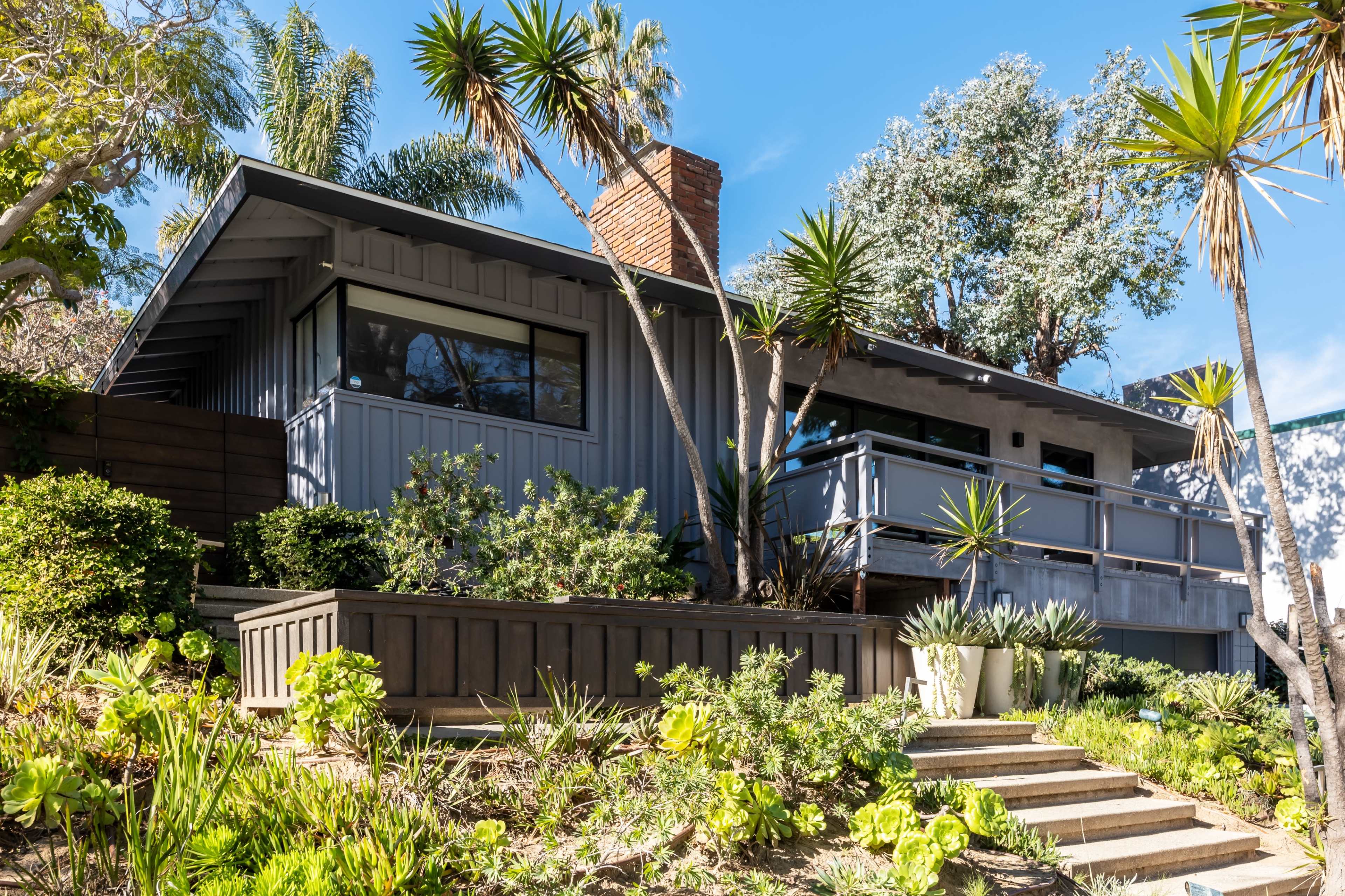 A modern house with a gray exterior and large windows is surrounded by lush greenery and landscaping on a hillside.