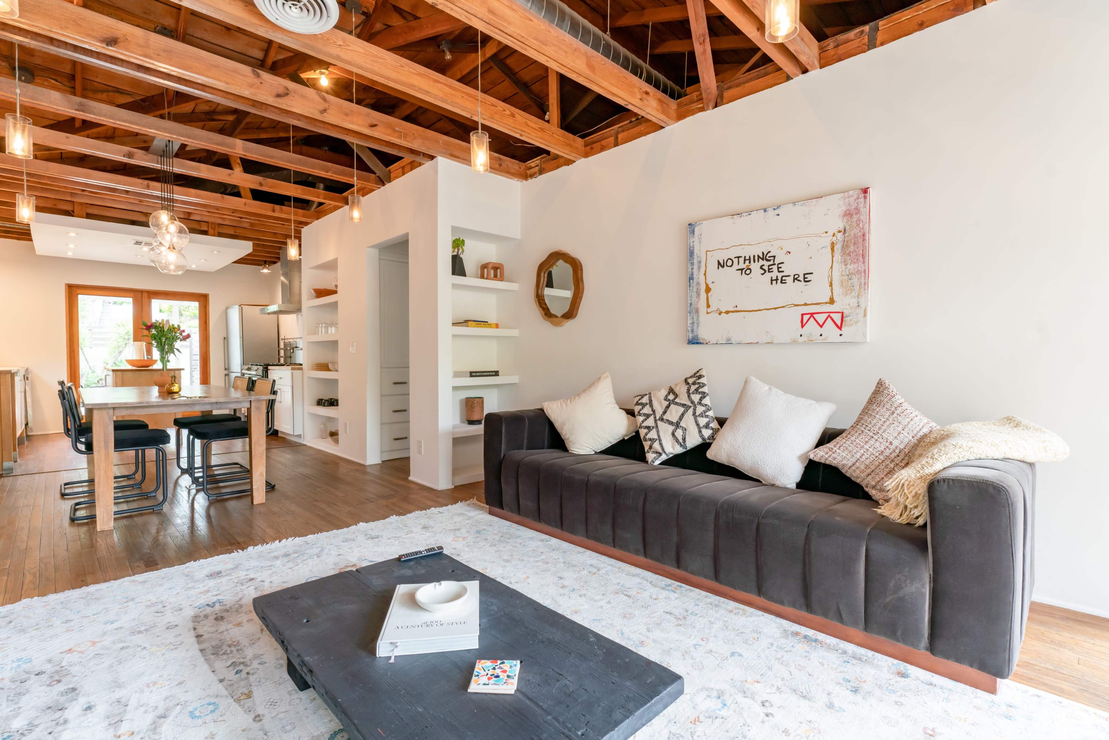 The image shows a modern living room with a dark sofa, a coffee table, and a wall art piece that reads "Nothing to See Here," featuring exposed wooden beams and open shelving.