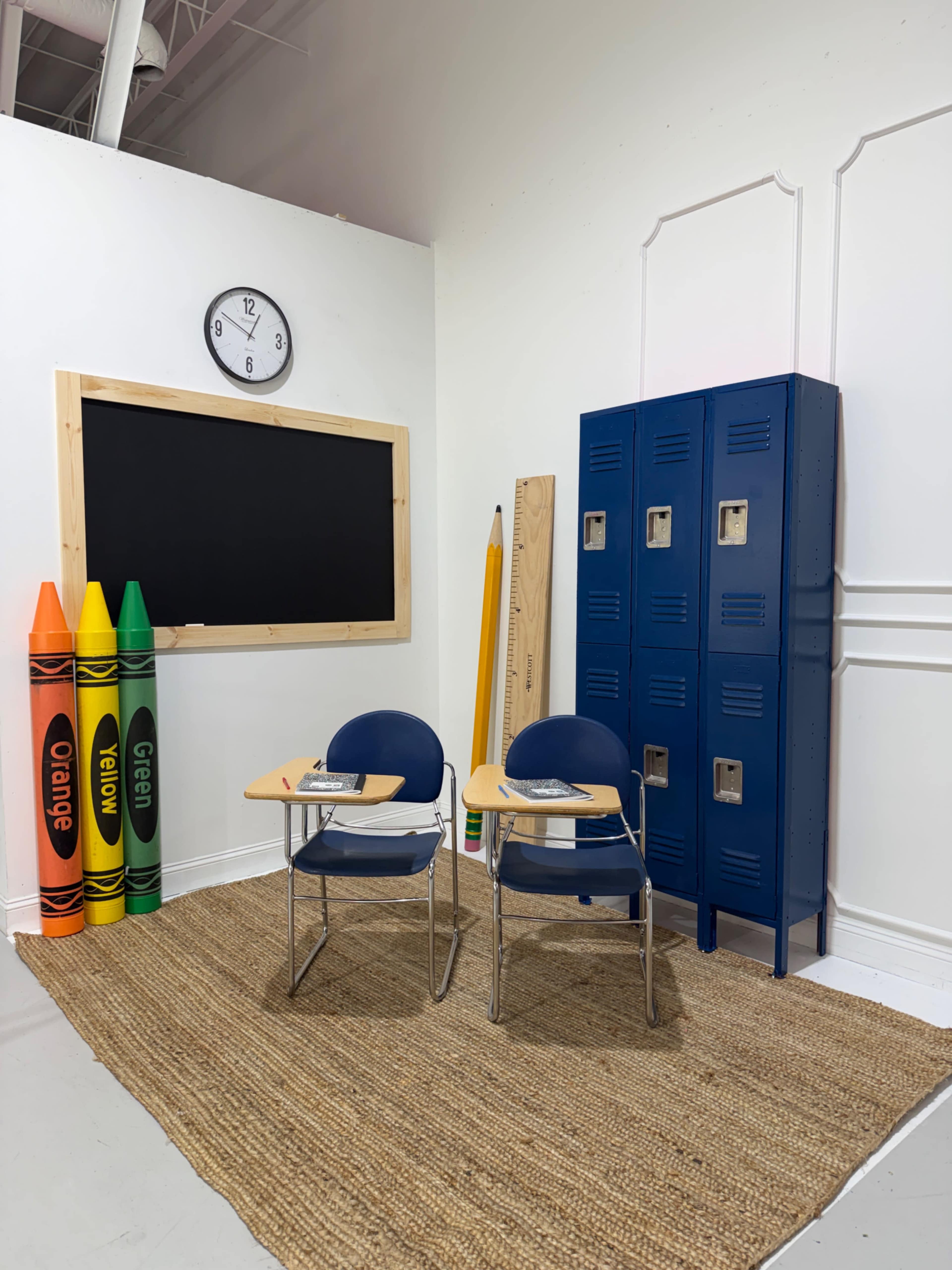 The scene features a classroom corner with two blue chairs and desks beside a chalkboard, colorful crayons, a large pencil, and a set of blue lockers on a woven rug.