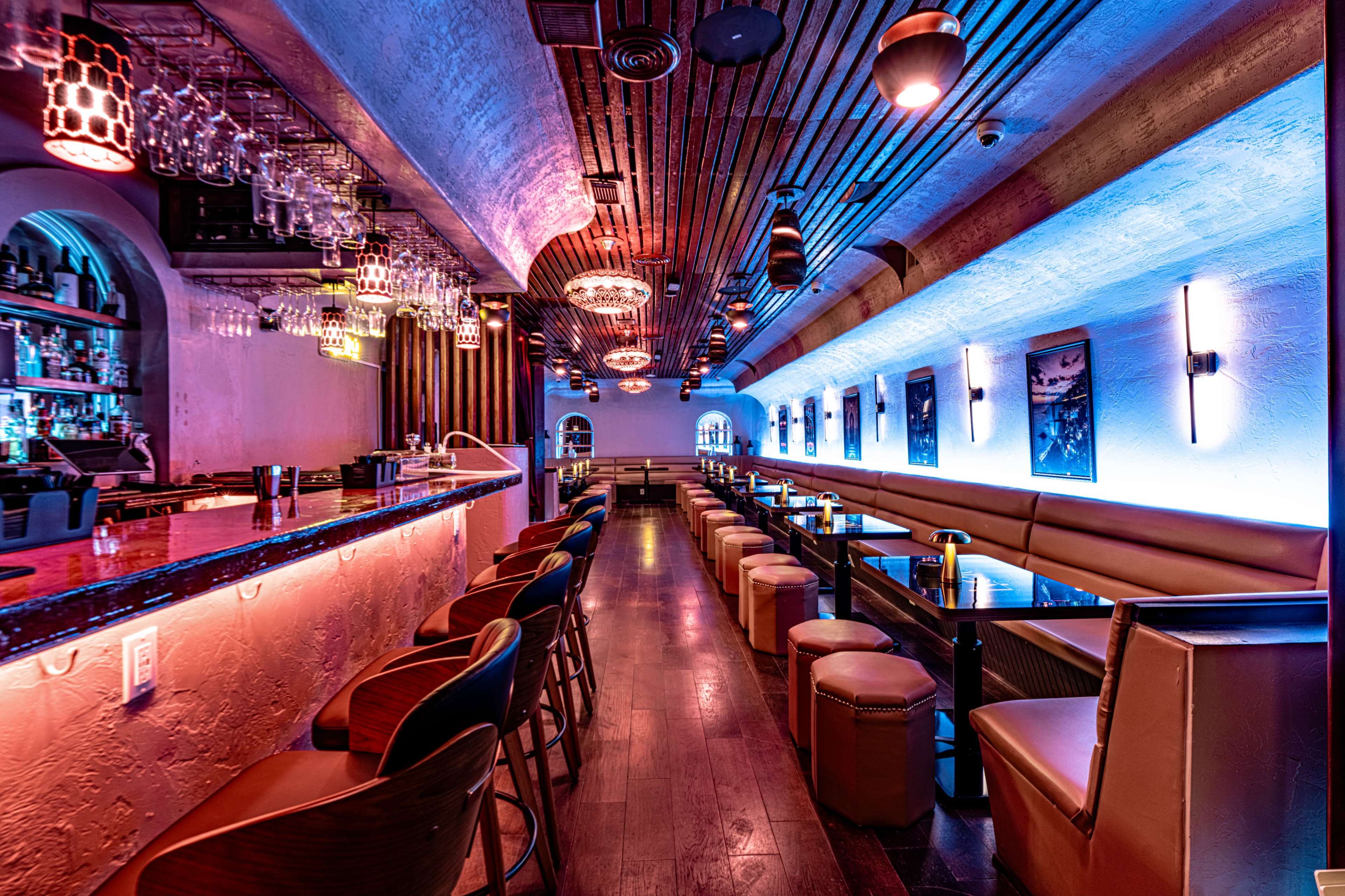 The image shows a modern bar interior with colorful lighting, high stools, and a long counter lined with glassware.