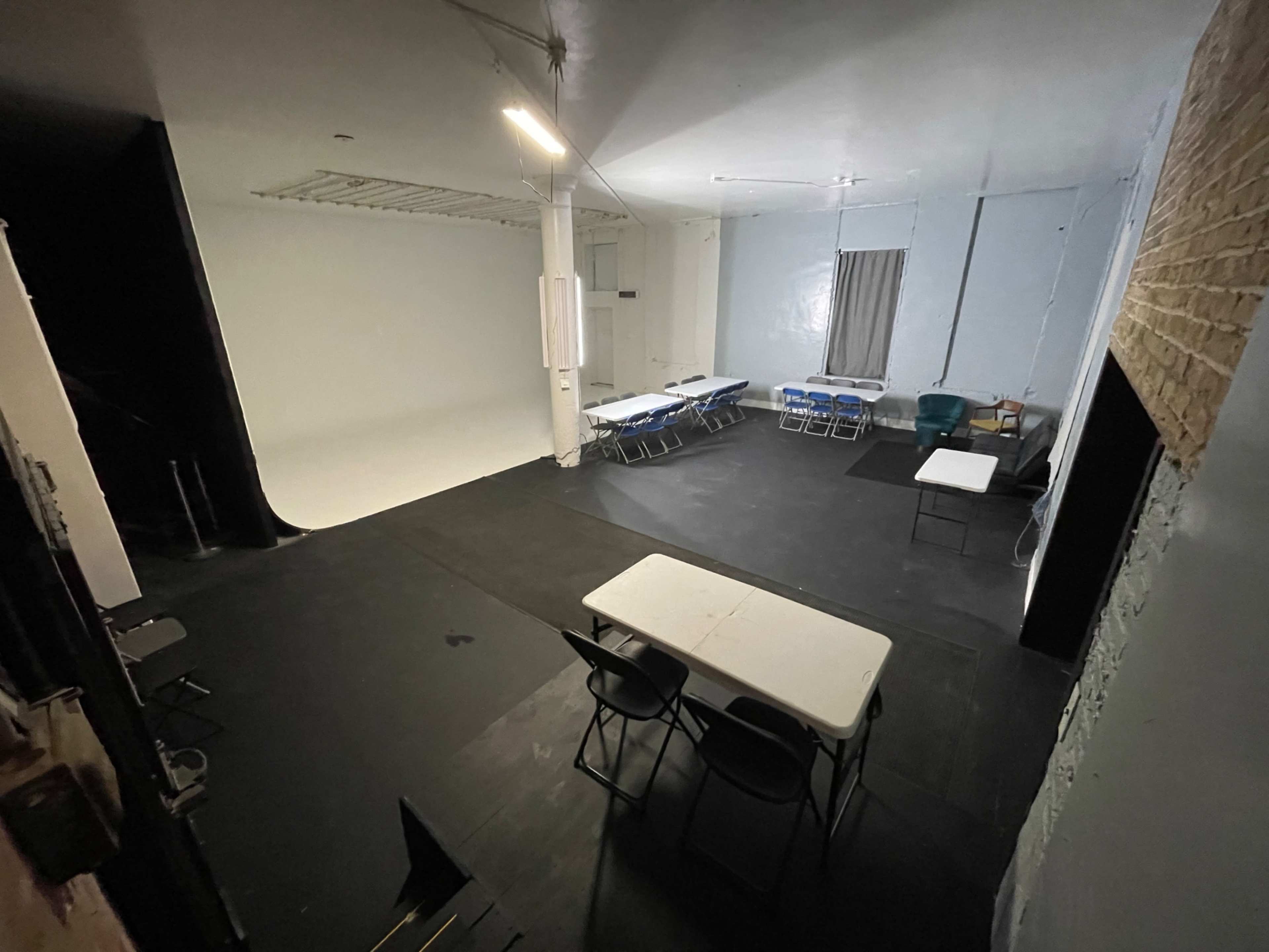 The image shows a spacious studio with a black floor, featuring a white backdrop, several tables and chairs arranged in two different sections, and a large window with a curtain.
