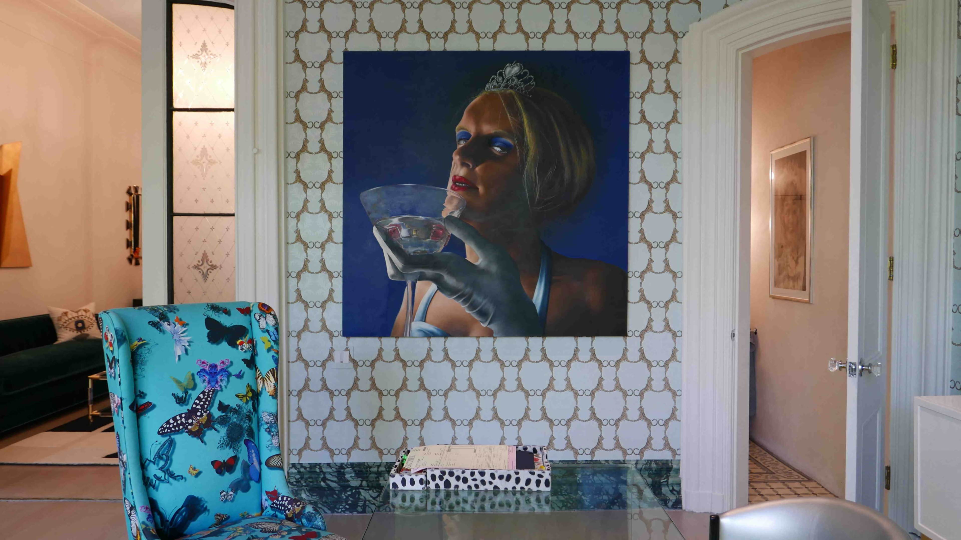 The image shows a brightly decorated room featuring a painting of a woman holding a drink on the wall, with a butterfly-patterned chair in the foreground.