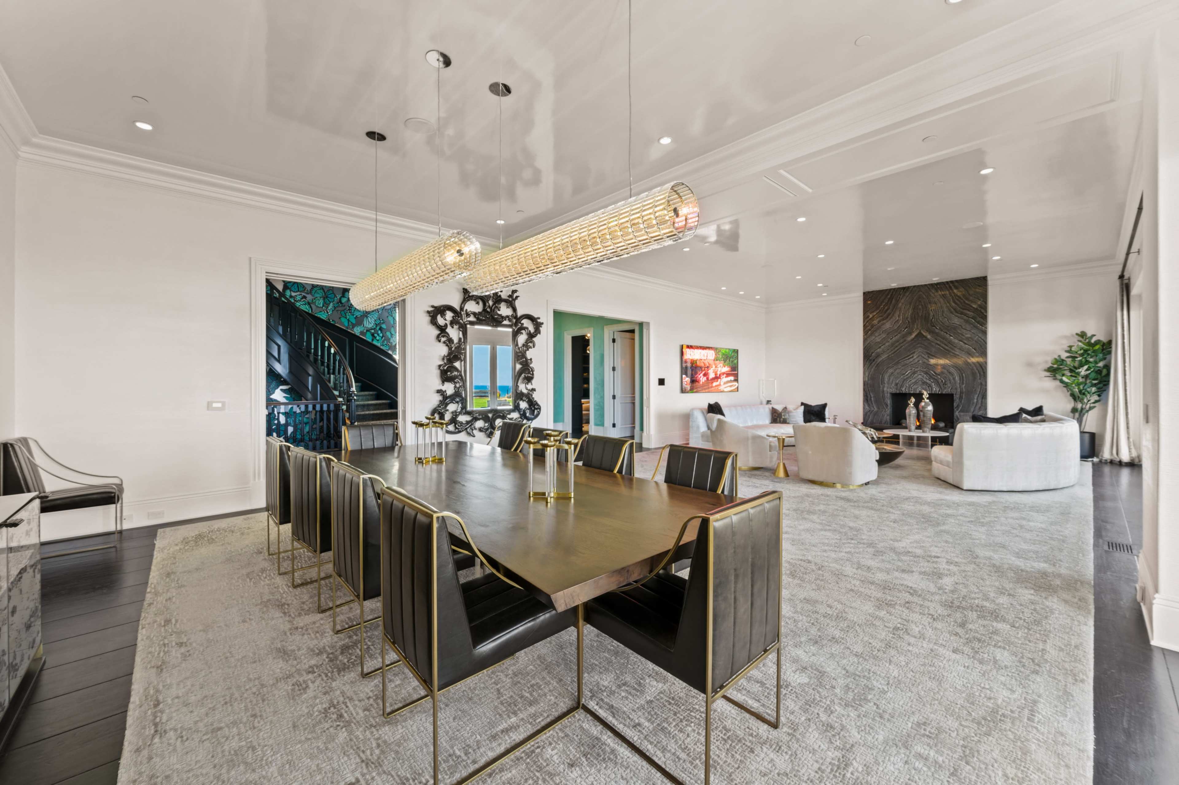 A spacious dining room features a long table with black chairs, illuminated by modern pendant lights, and opens to a living area with a large window providing a view.
