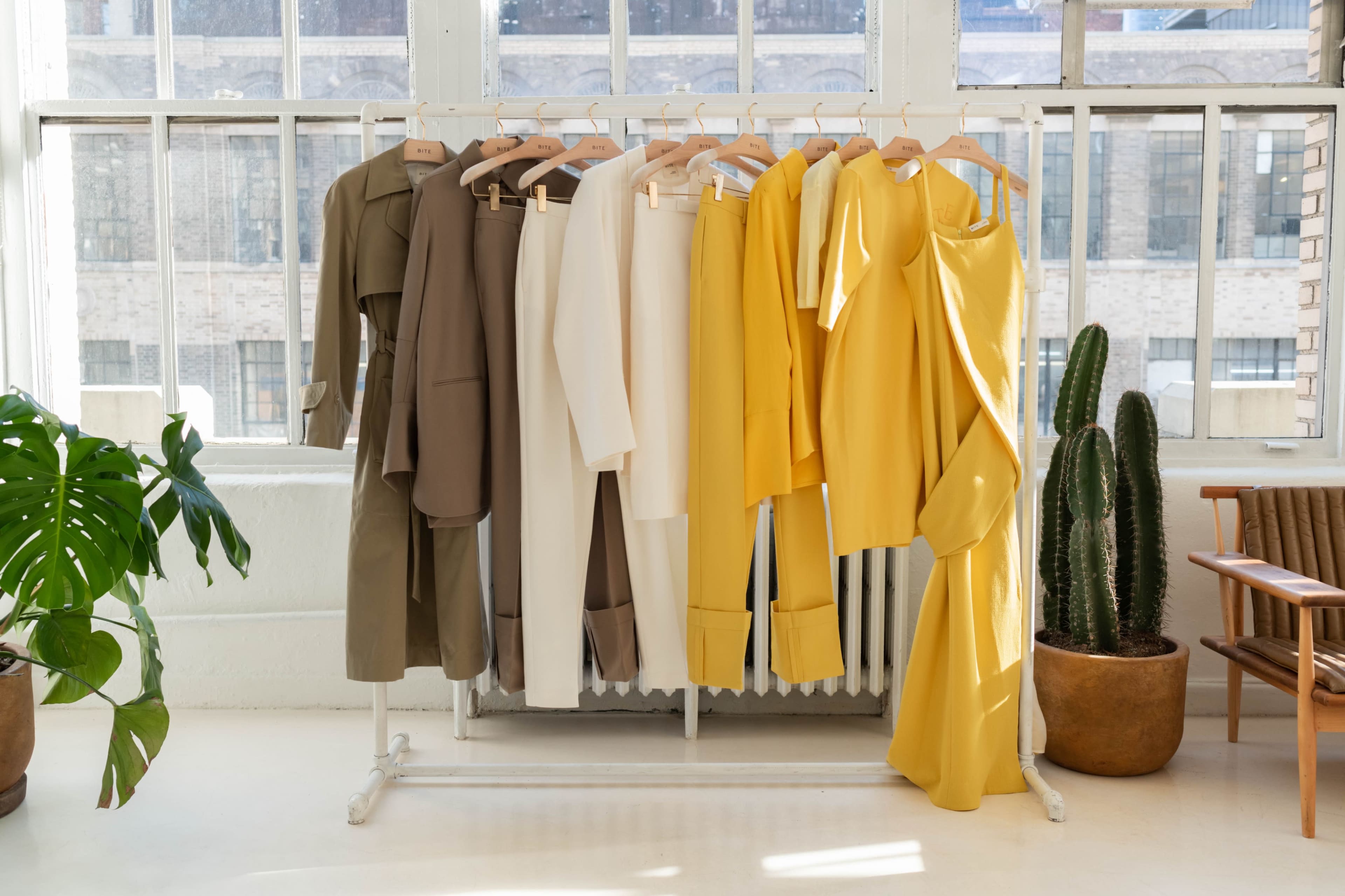 A rack displays a collection of clothing in neutral and yellow tones, positioned near large windows with a view of a plant and a chair in the background.