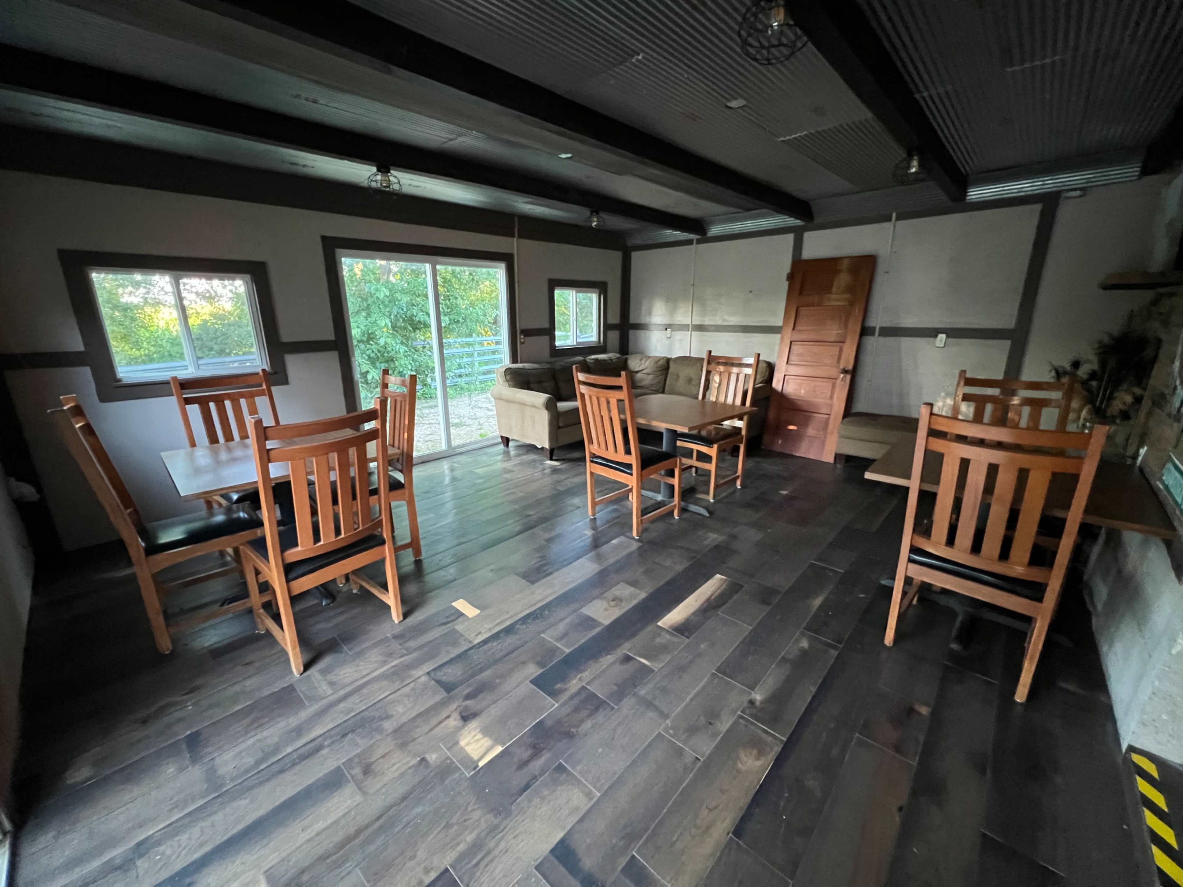 The image shows a spacious room with wooden flooring, several wooden dining tables and chairs, a couch, and a large sliding glass door leading outside.