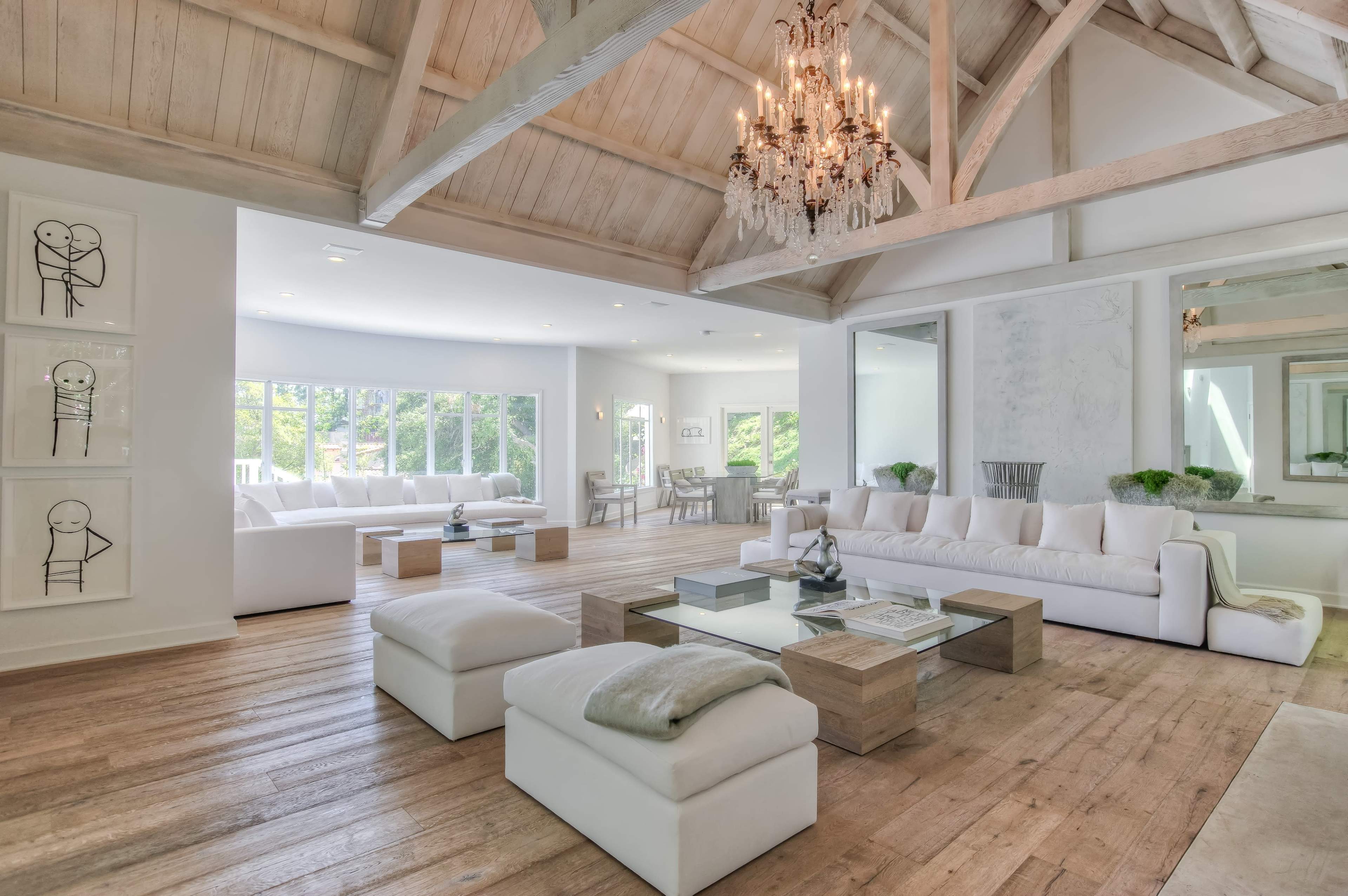 A spacious and bright living room features high wooden ceilings, large windows, and minimalist furniture arranged around a central coffee table.
