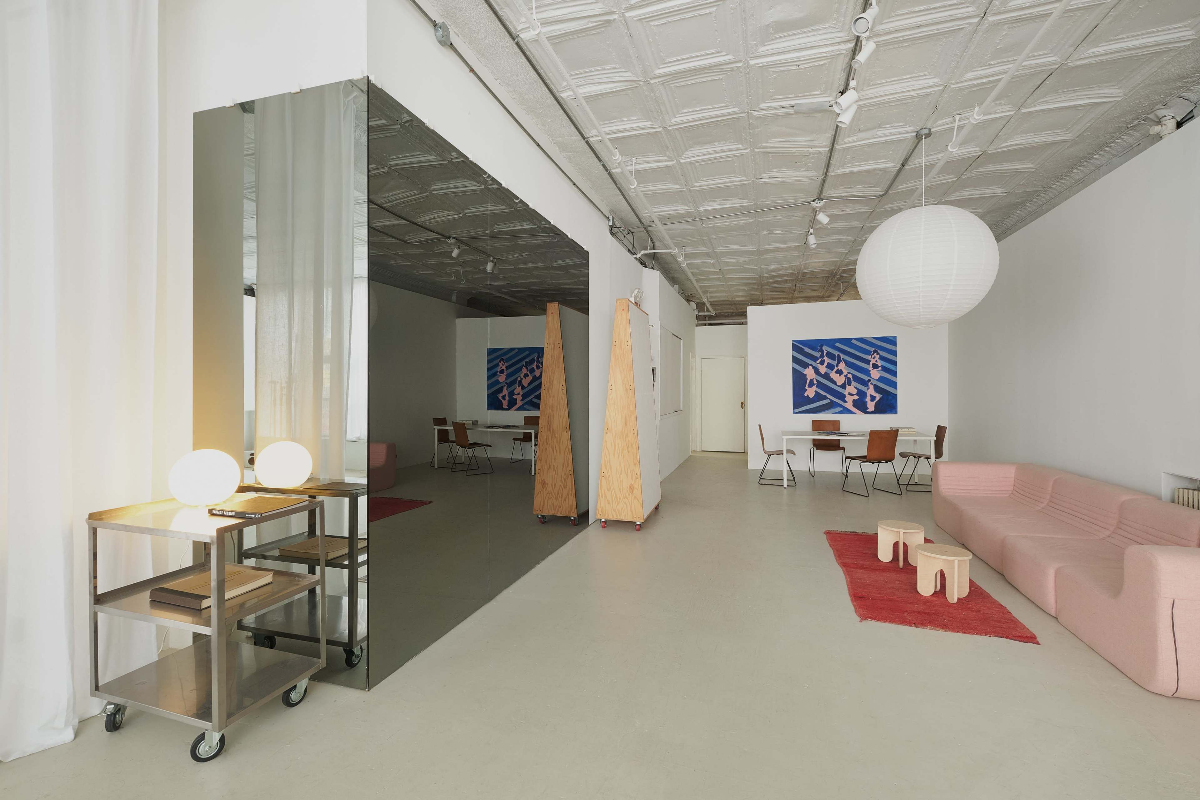 The image depicts a modern, minimalist interior space featuring a pink sofa, a mirrored wall, wooden dividers, and a dining area with a round paper lantern hanging from the ceiling.