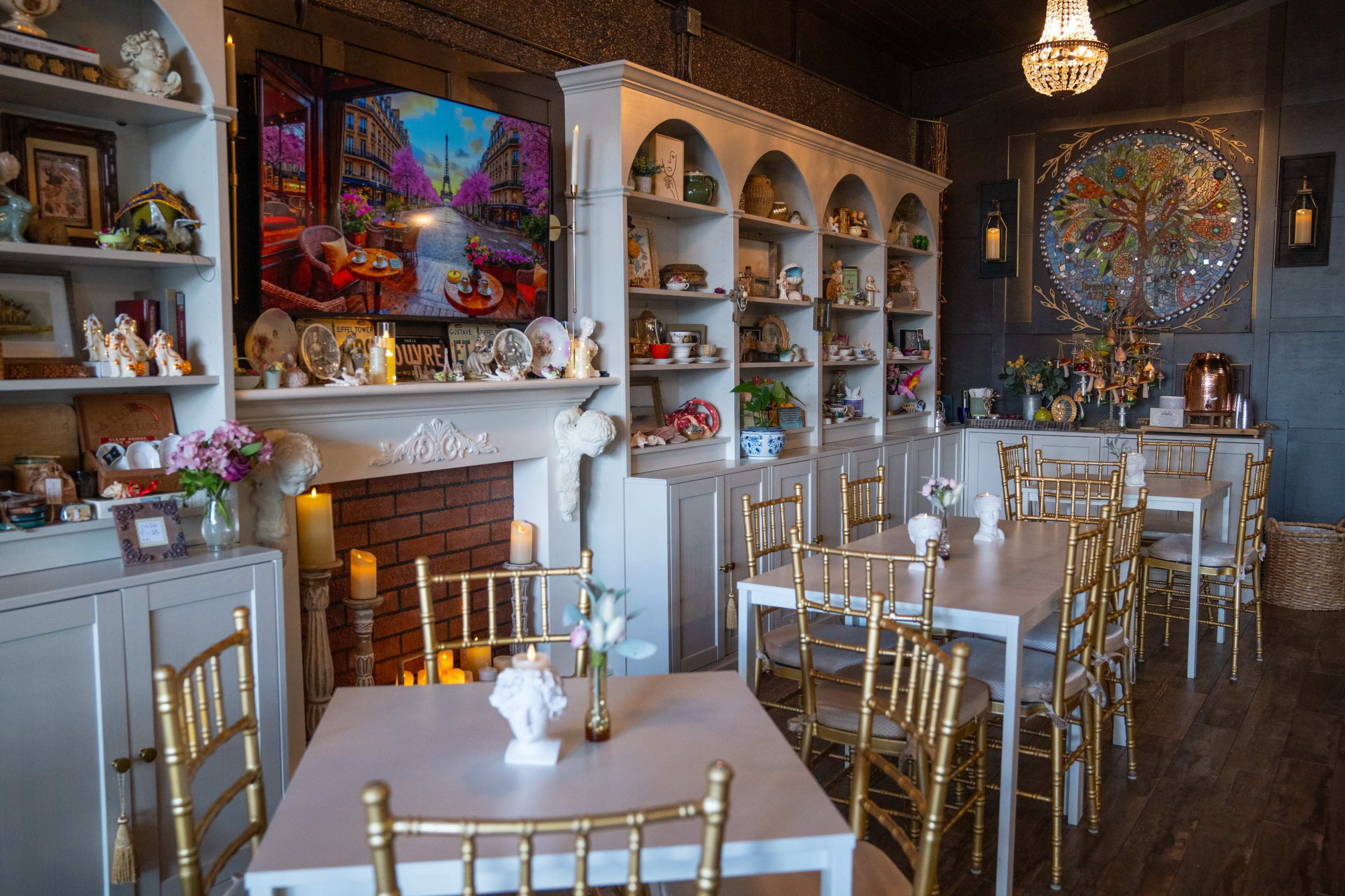 The image shows a quaint interior of a café with white tables and gold chairs, shelves filled with various decorative items, and a colorful painting on the wall.