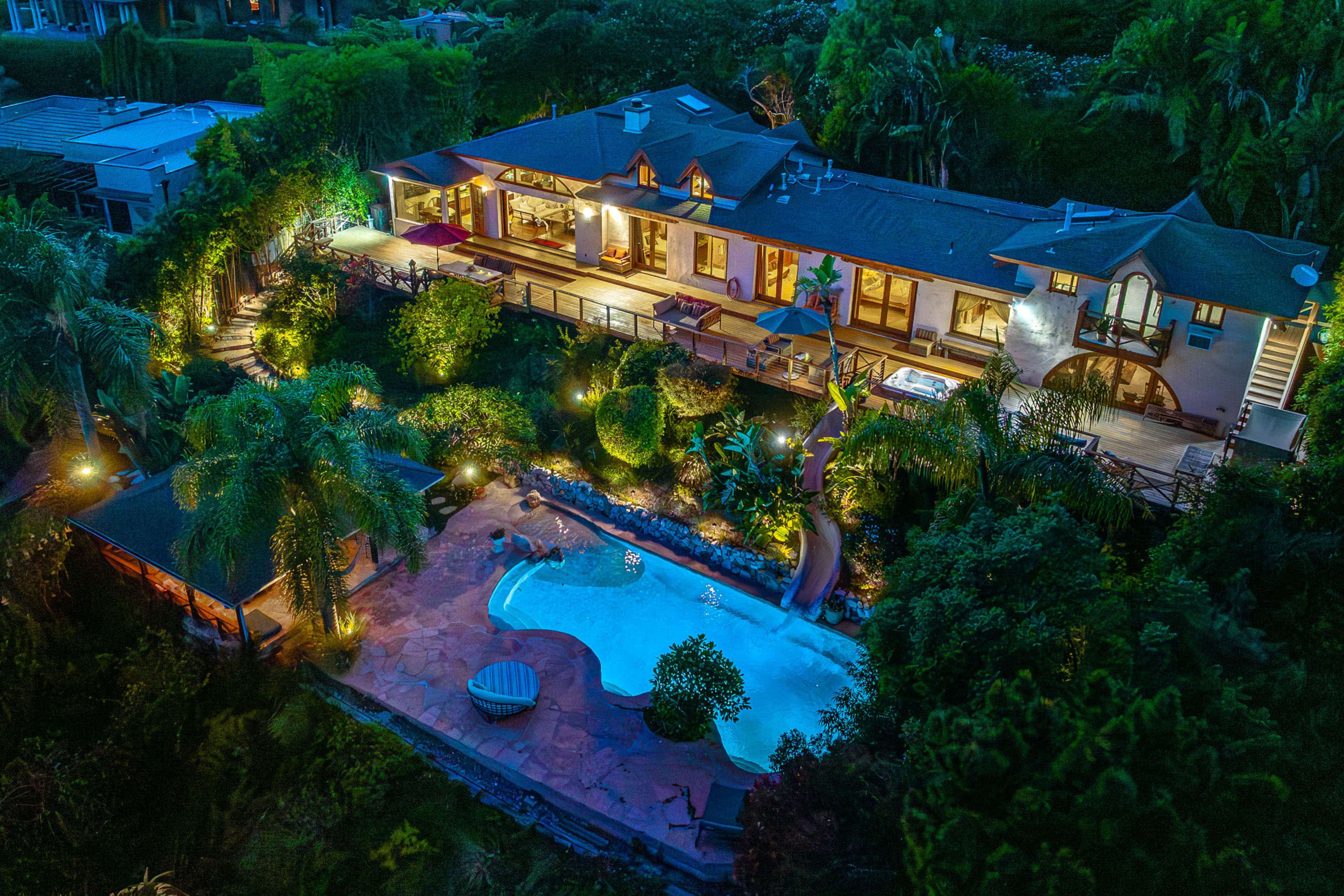 A large house surrounded by lush greenery, an illuminated swimming pool, and a slide, all set against a darkening evening sky.