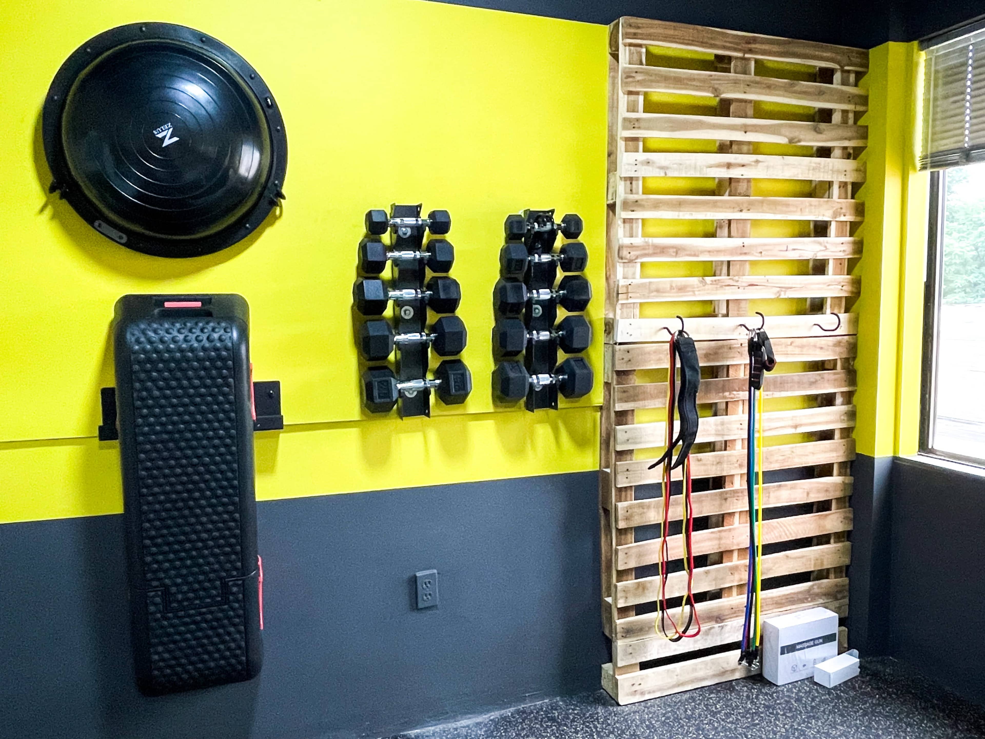 A fitness corner with exercise equipment including weights, resistance bands, a balance board, and a wooden pallet for storage, set against a yellow and gray wall.