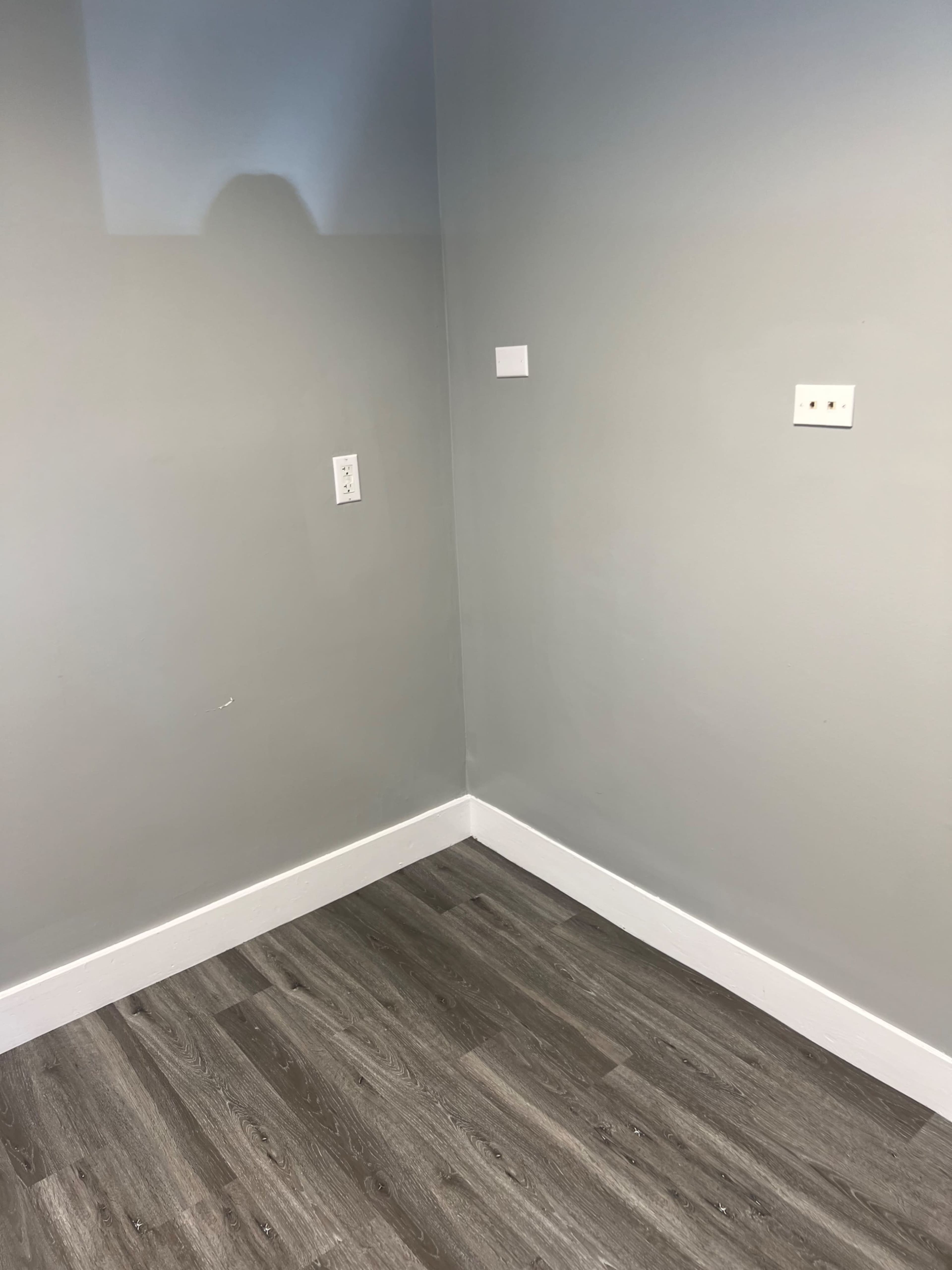 A corner of a room with gray walls and a wooden floor, featuring two electrical outlets on the wall.
