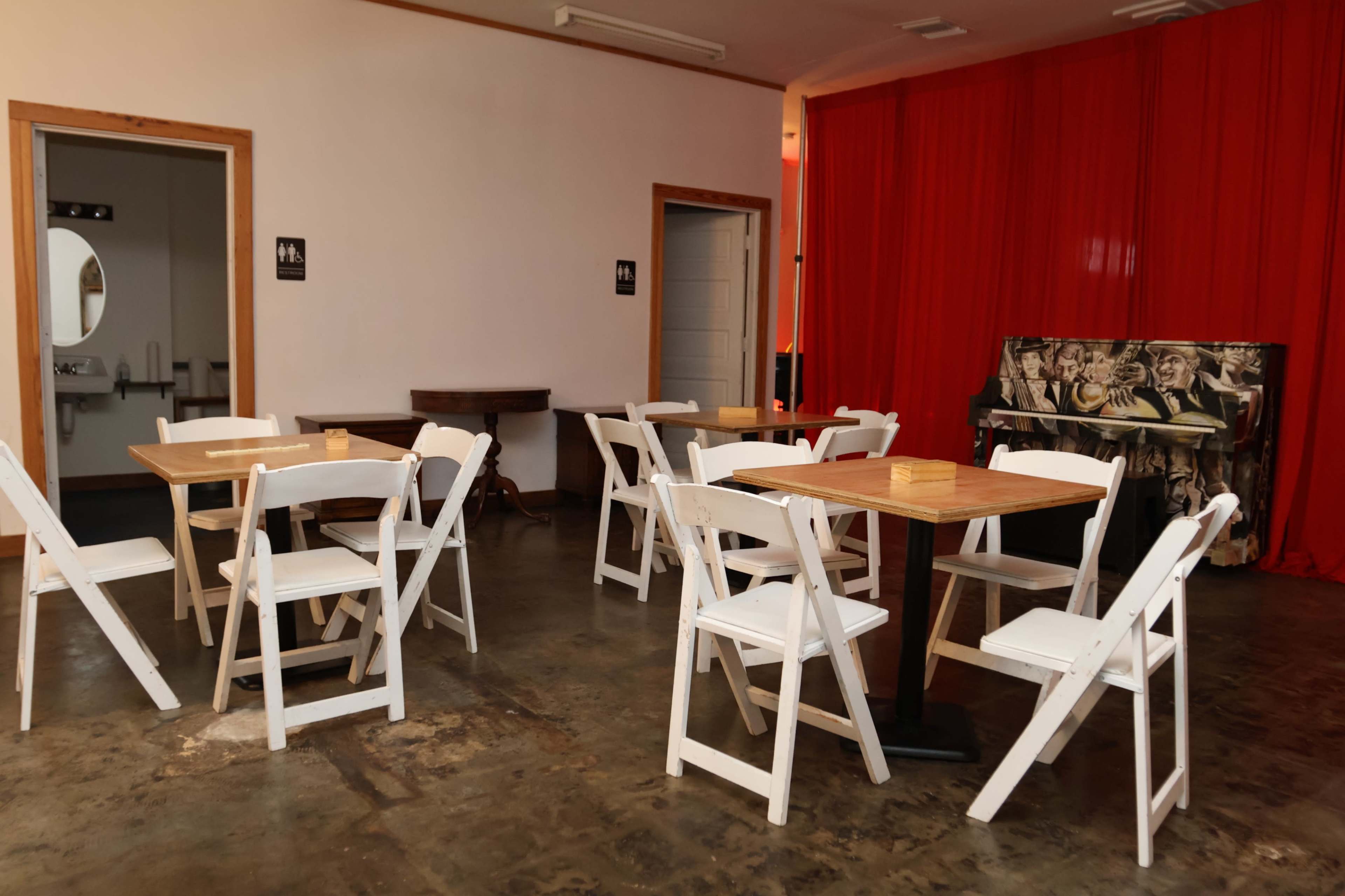 The image shows a room with several white folding chairs and tables arranged around a dark floor, with a red curtain and a painted piano in the background.