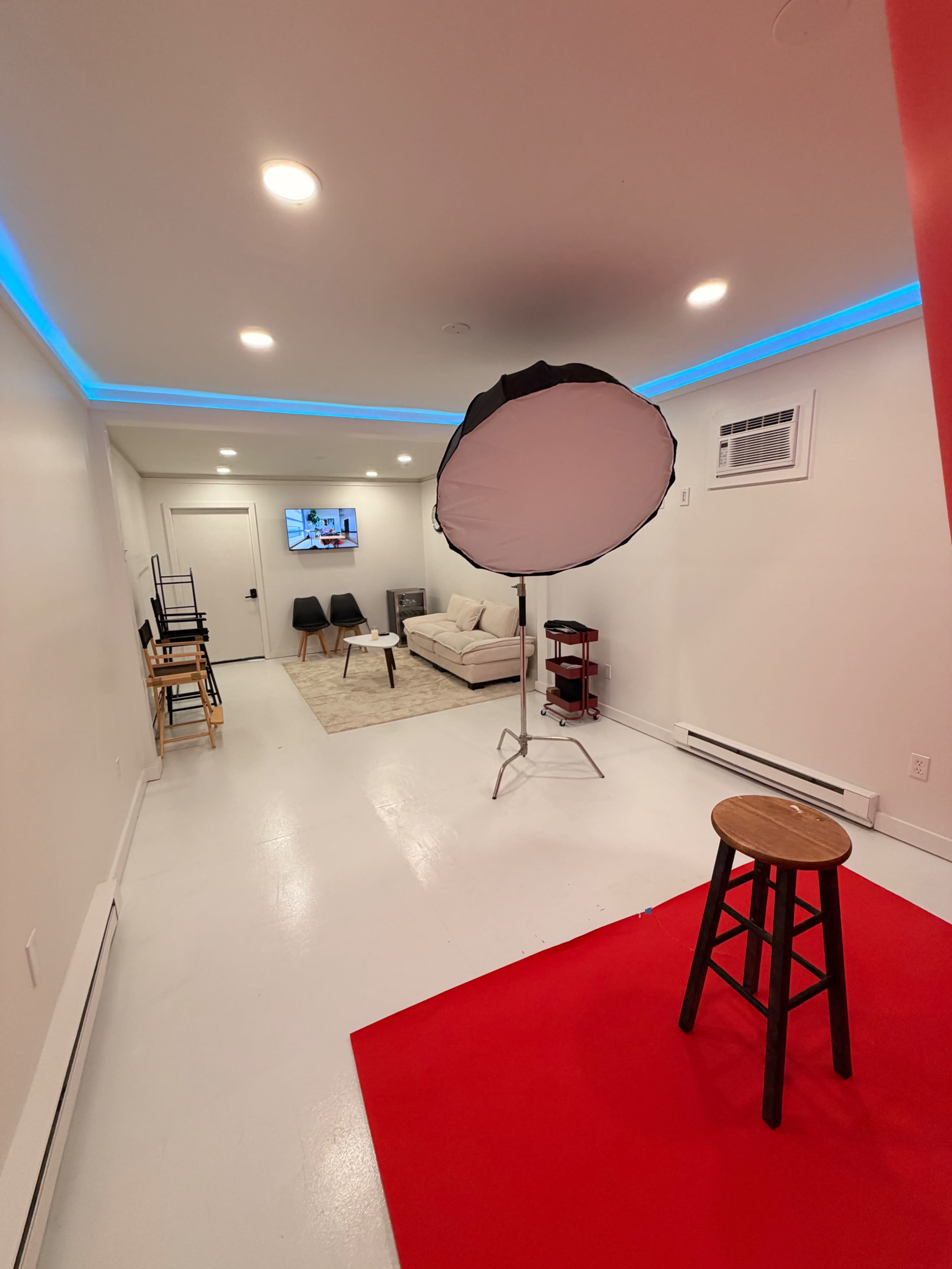 The image shows a well-lit studio space featuring a red carpet, a stool, a large softbox light, a white couch, and a TV mounted on the wall.