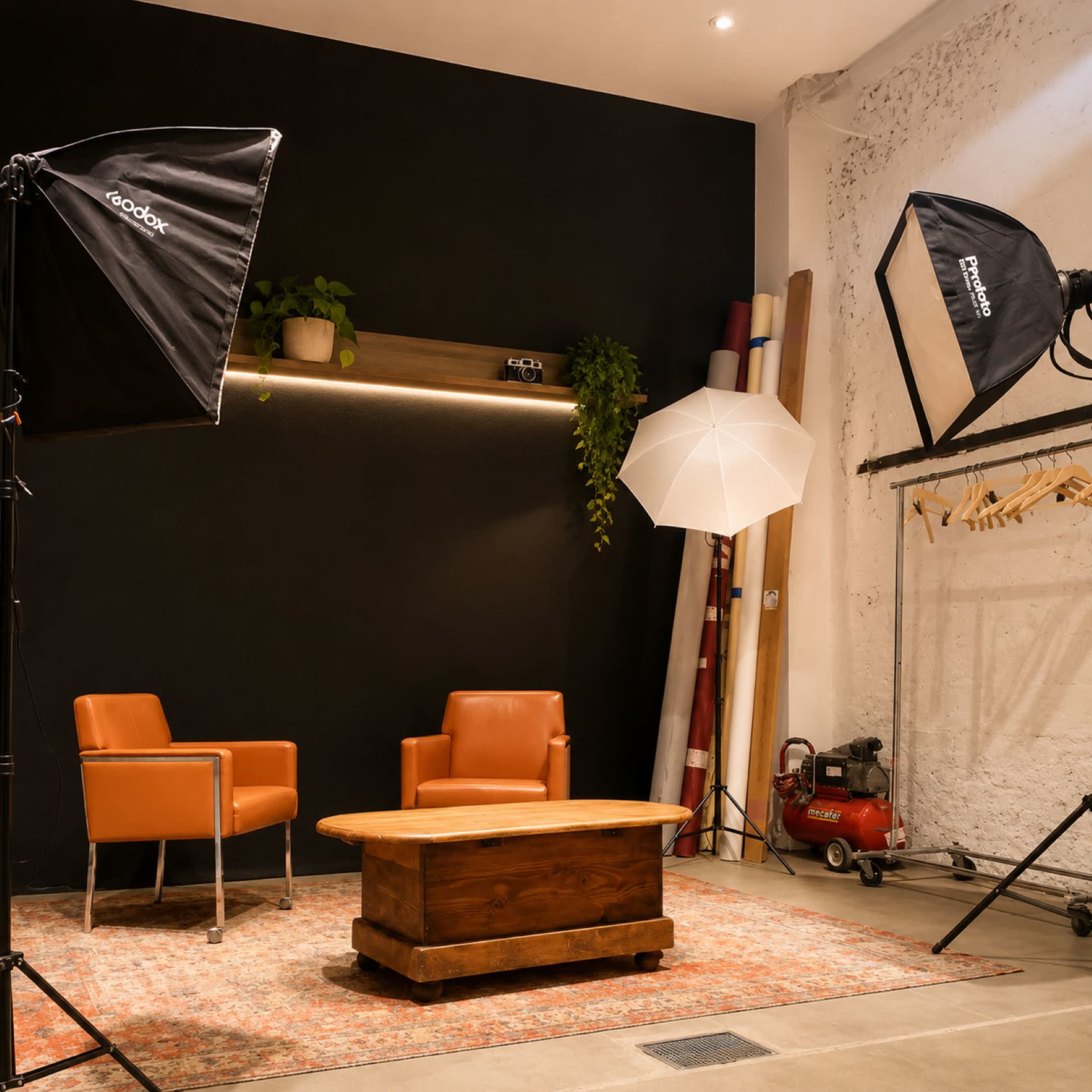 The image shows a photo studio setup featuring two orange chairs, a wooden coffee table, and professional lighting equipment against a black wall.