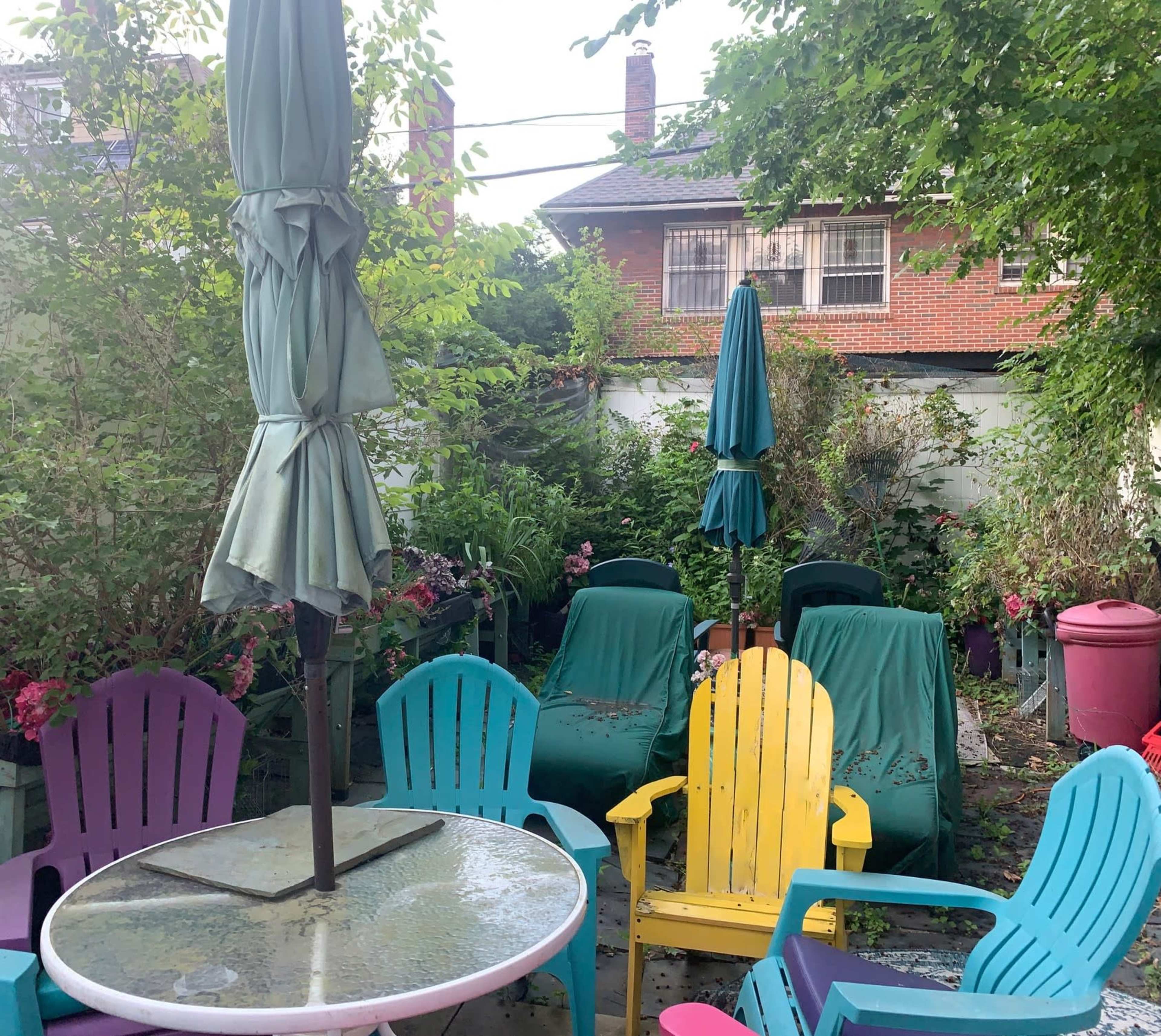 The image shows a small outdoor seating area with colorful chairs, a table, and several umbrellas surrounded by greenery and plants.