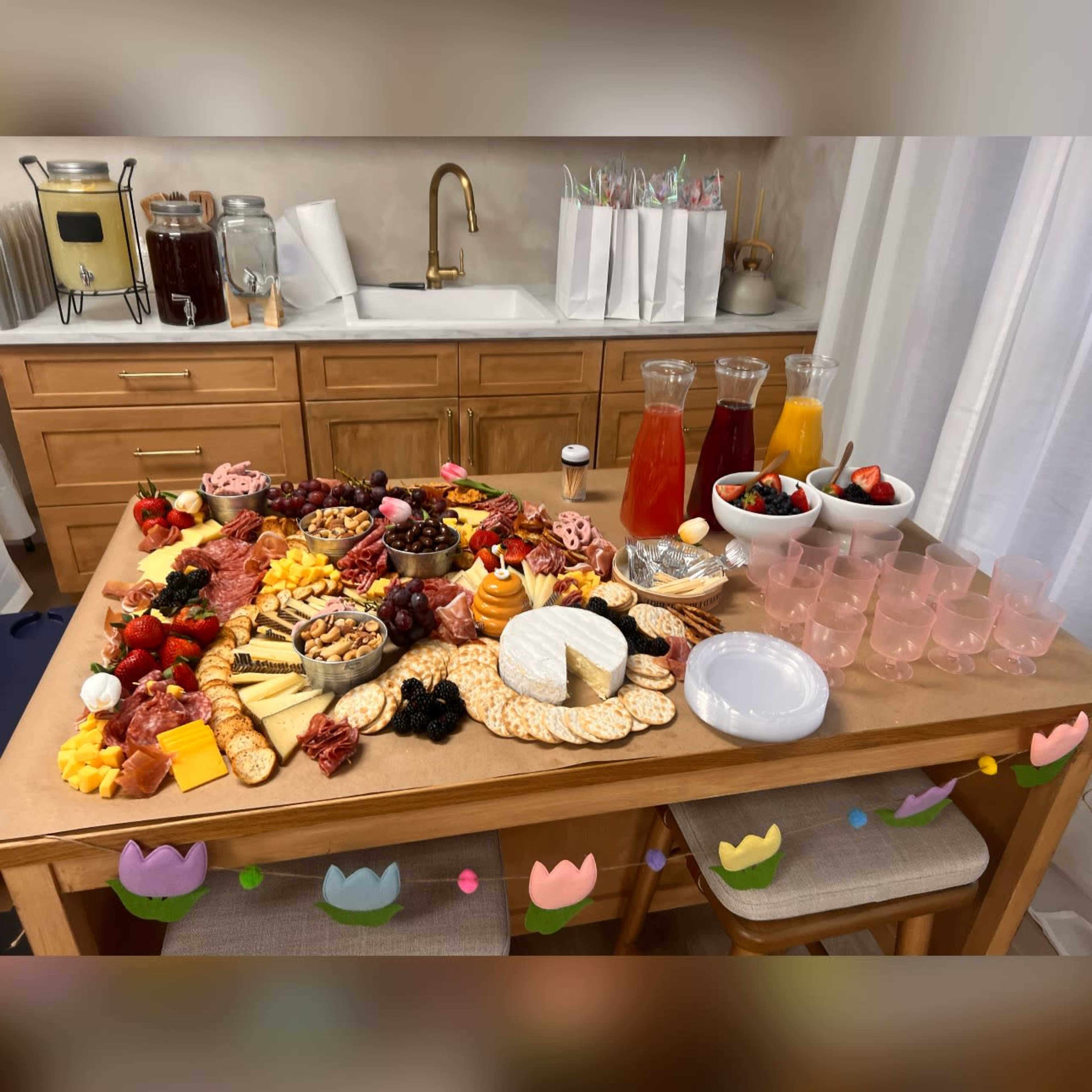 A wooden table is spread with an elaborate assortment of cheeses, meats, fruits, nuts, crackers, and beverages in glass pitchers and bowls.