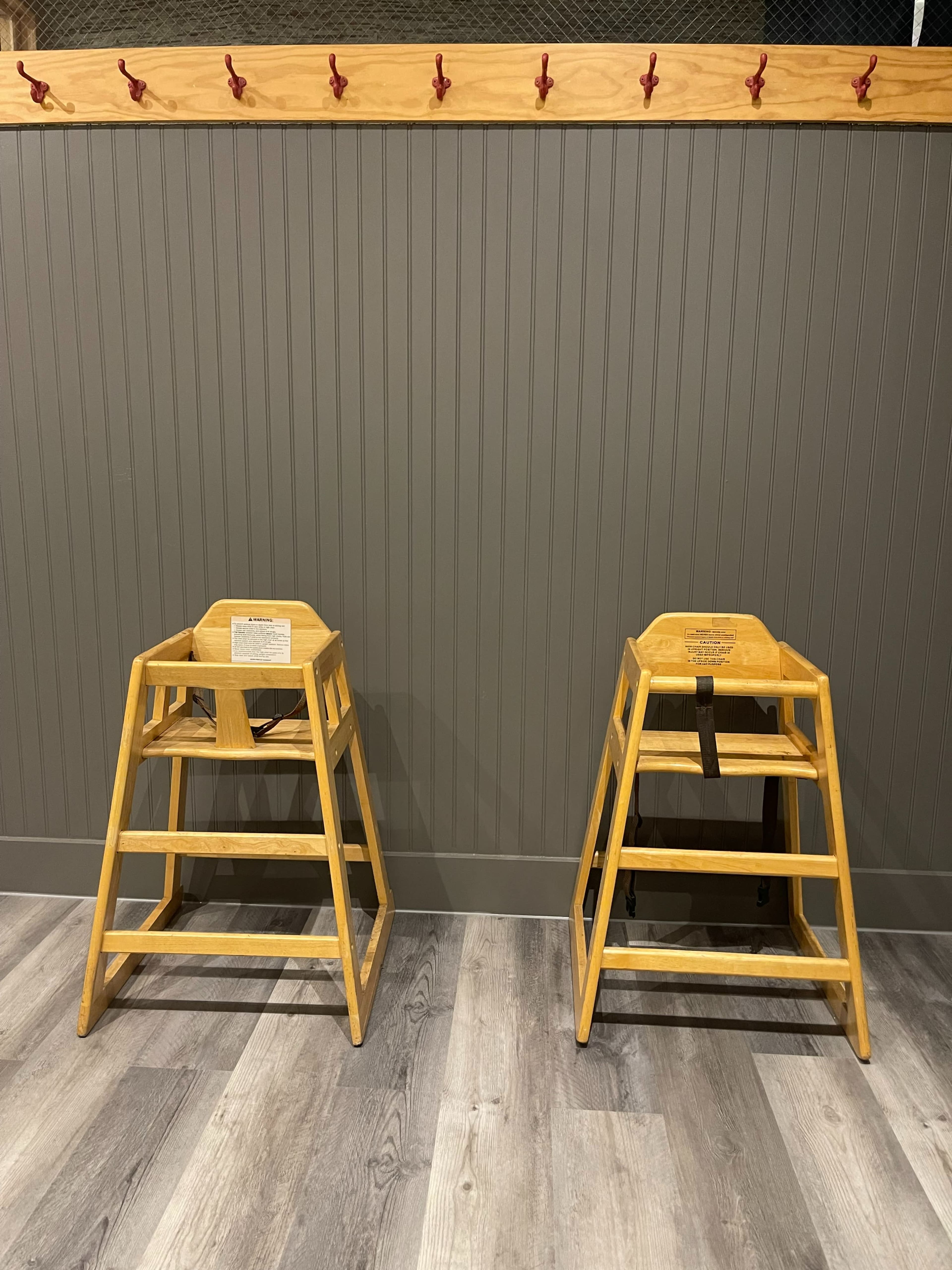 Two wooden high chairs are positioned against a gray paneled wall with a row of red hooks above them.