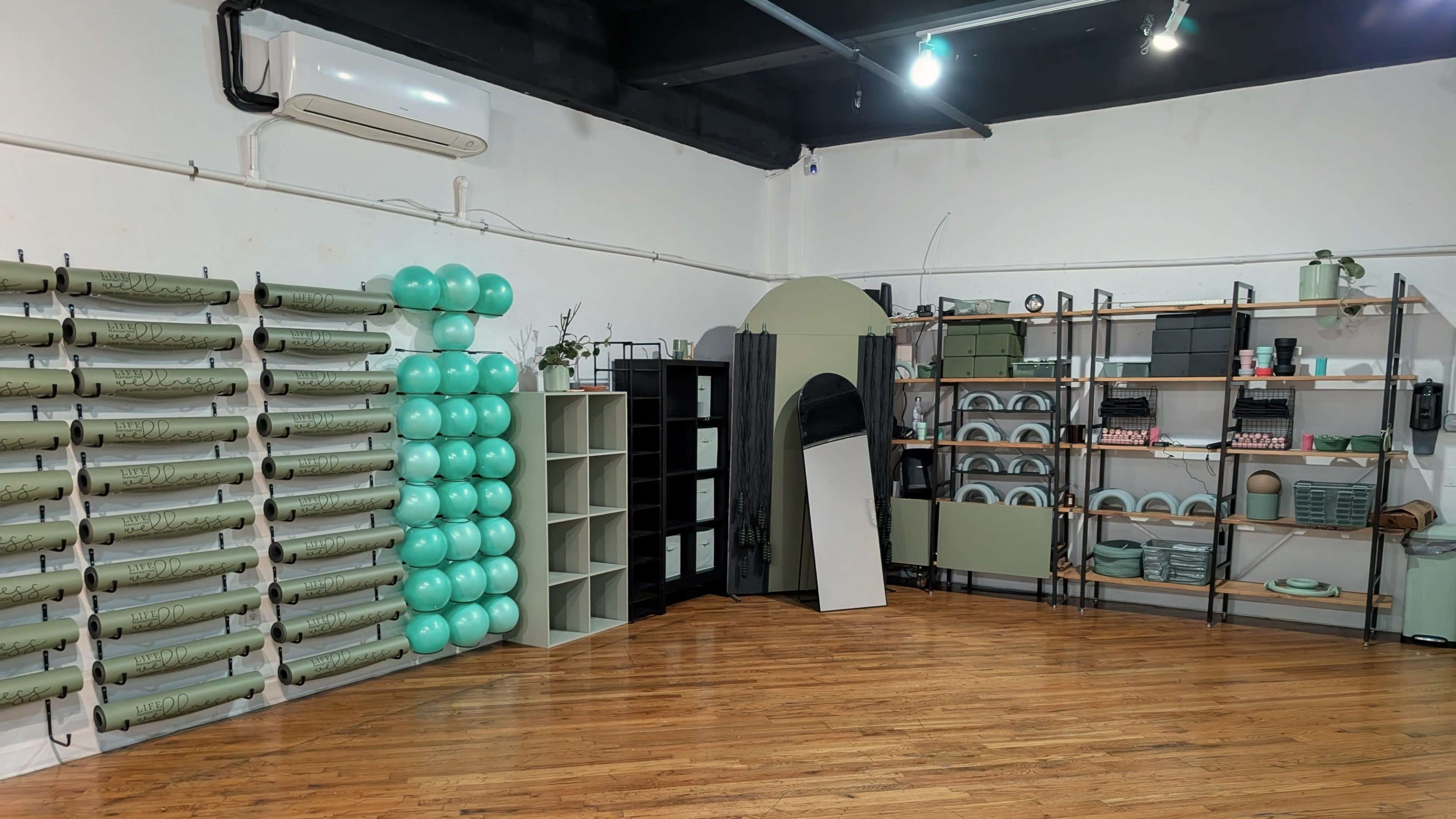 The image shows a spacious room with wooden flooring, featuring a wall of green rolls, a stack of turquoise balloons, a black shelving unit, and various shelves displaying household items.