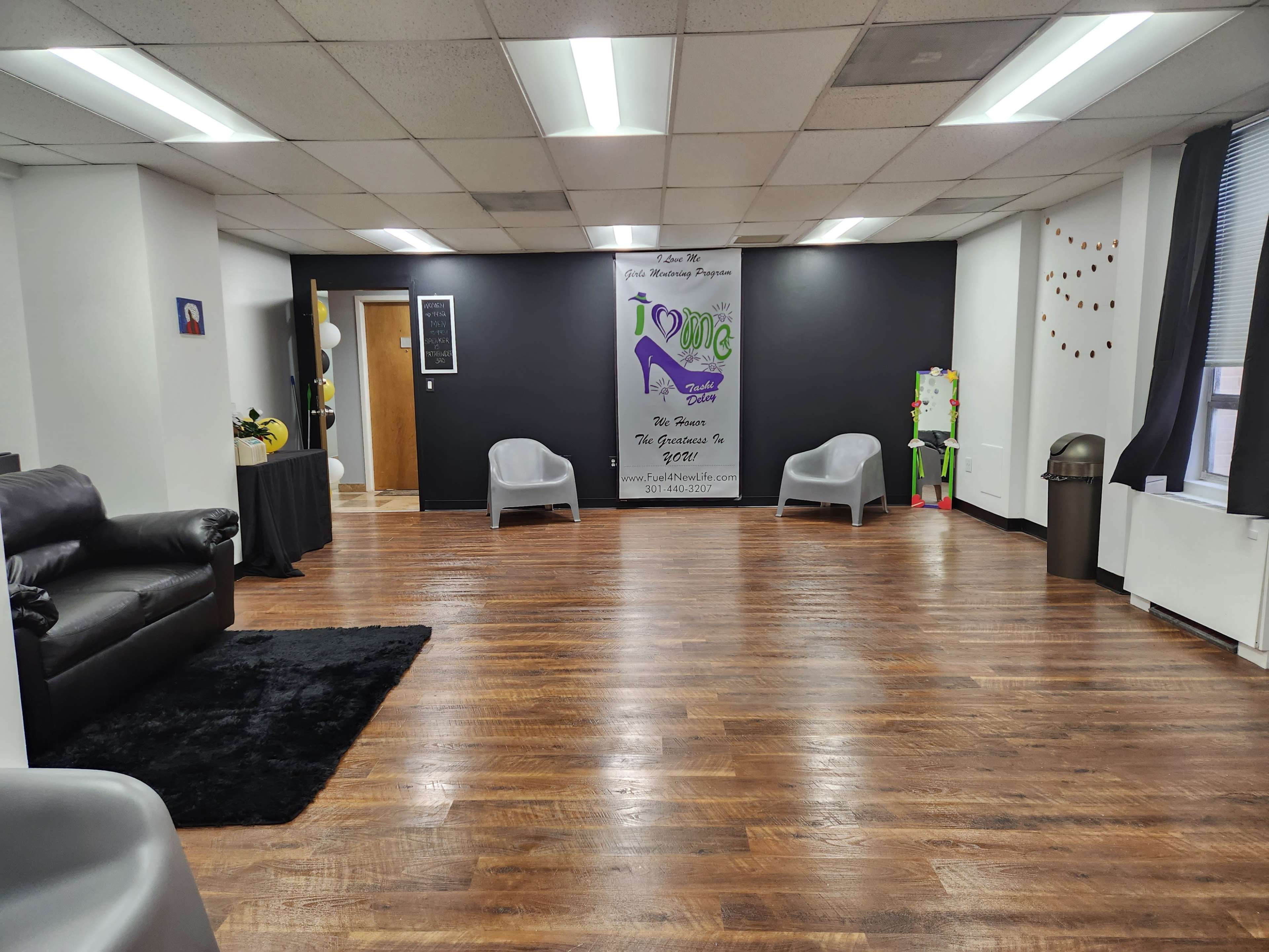 A spacious room with dark walls, hardwood flooring, and several seating areas, including two light gray chairs, a black couch, and a black table near the entrance.