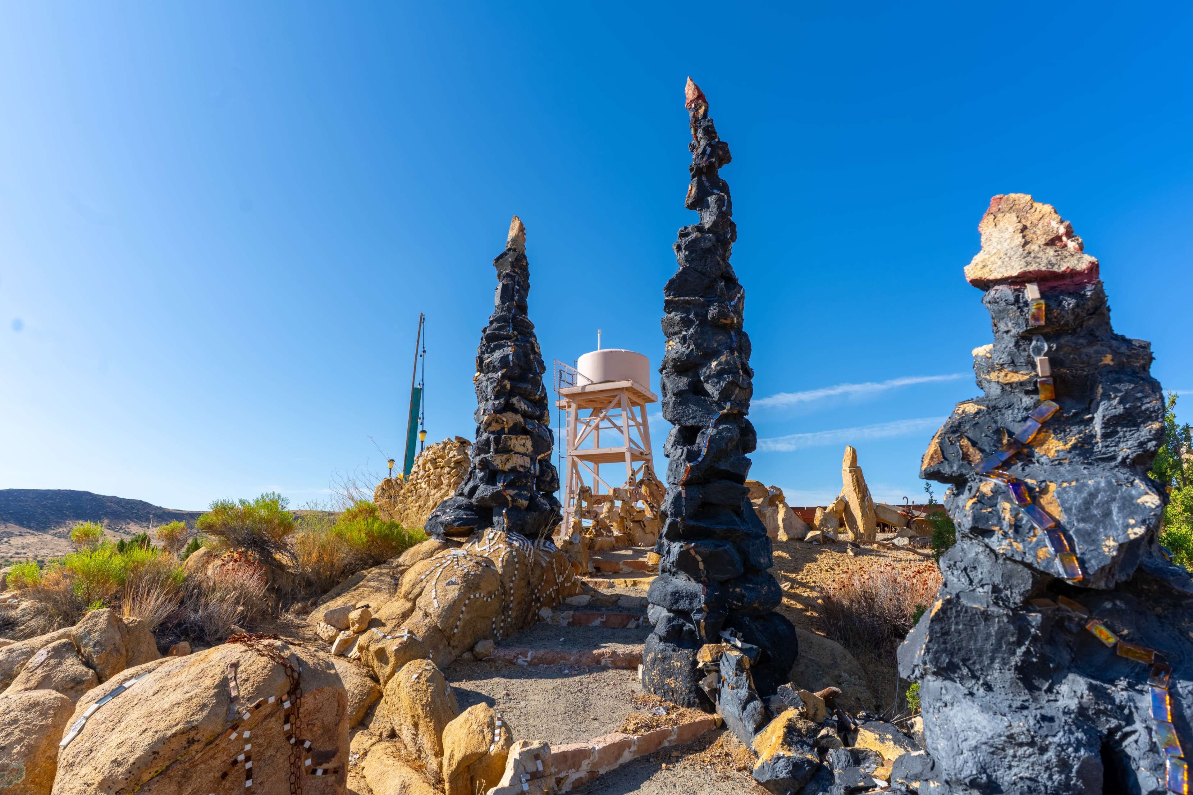 The image shows a rocky pathway flanked by tall, black stone formations leading towards a water tank on a wooden structure against a clear blue sky.