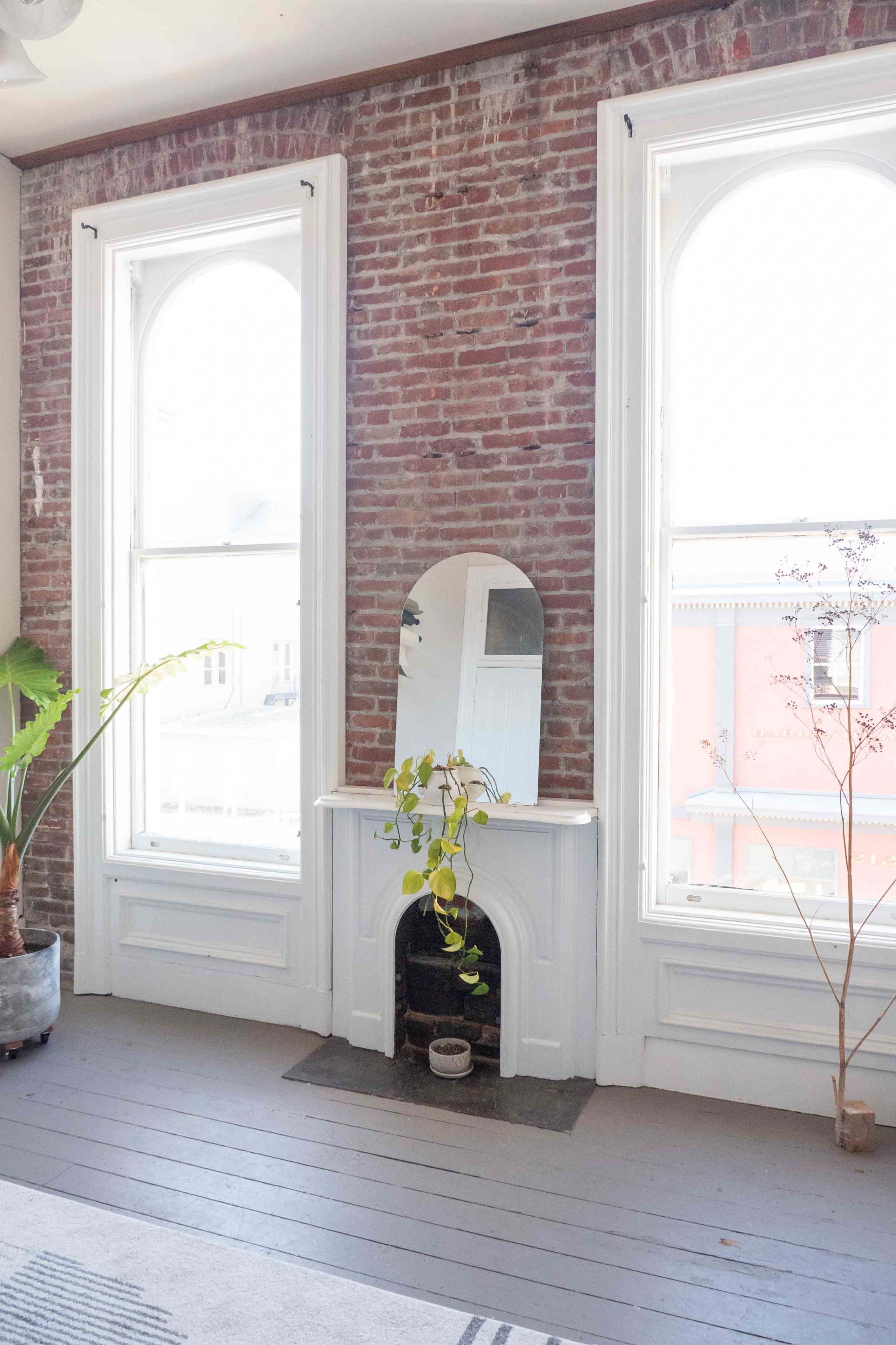 The image features a room with exposed brick walls, two large arched windows, and a mirror above a small fireplace surrounded by plants.