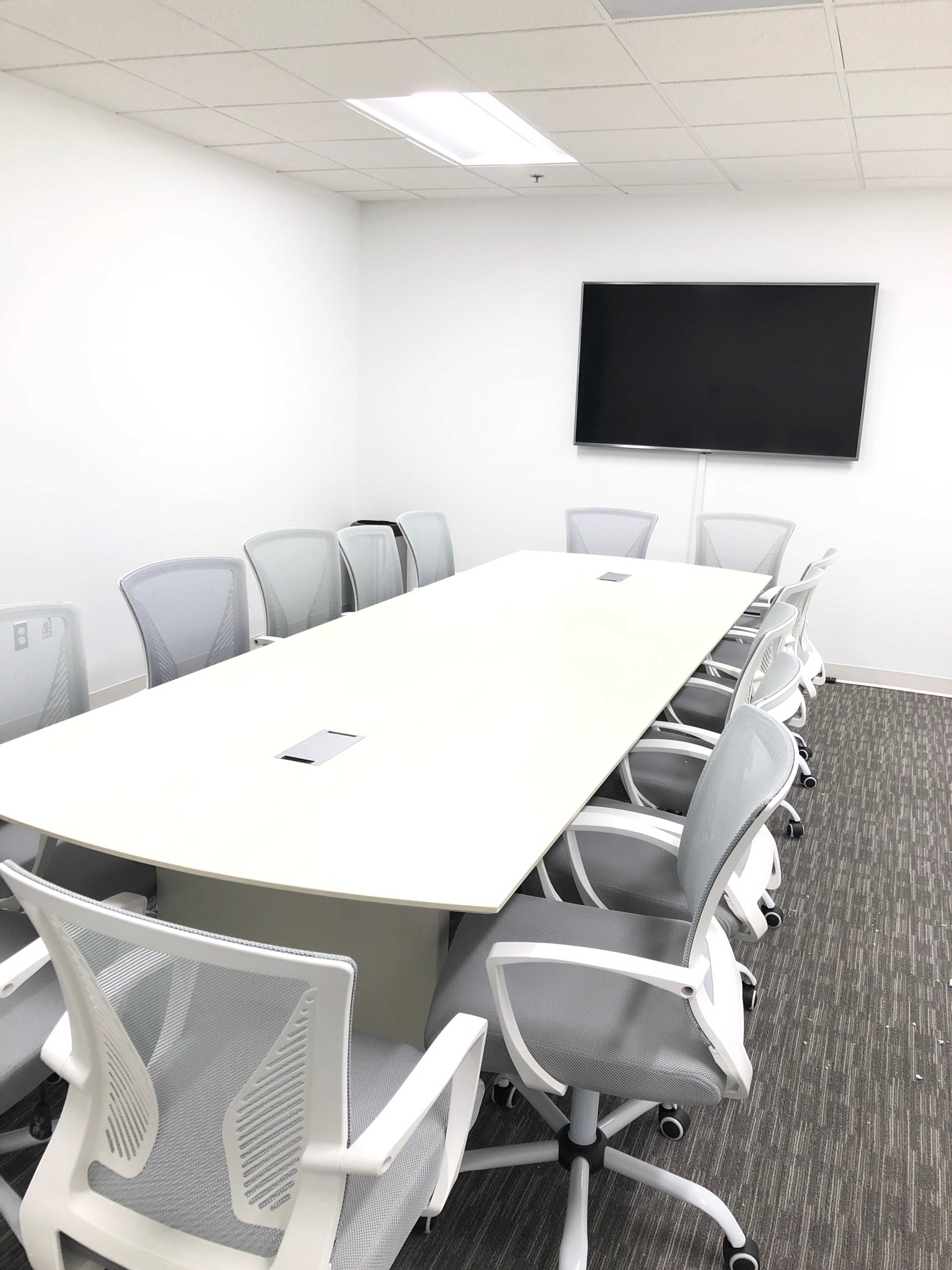 A modern conference room features a long white table surrounded by several white office chairs, with a large screen mounted on the wall.