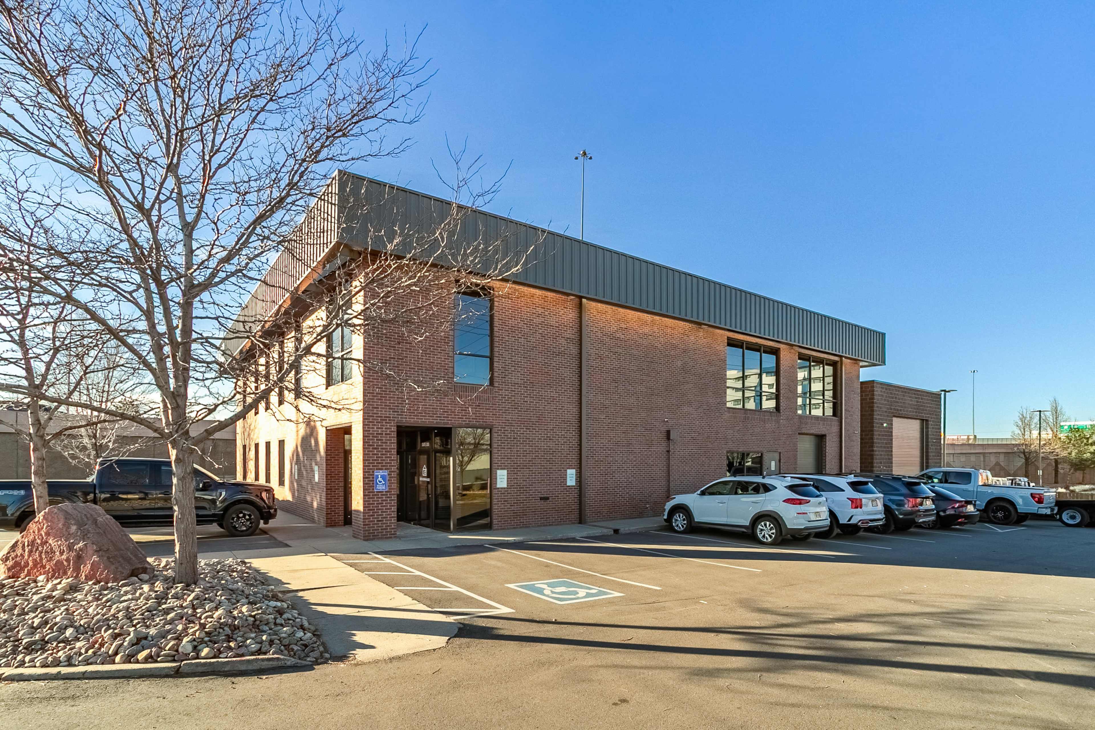 The image shows a brick commercial building with a metal roof, surrounded by a parking lot and a landscaped area with rocks and a barren tree.