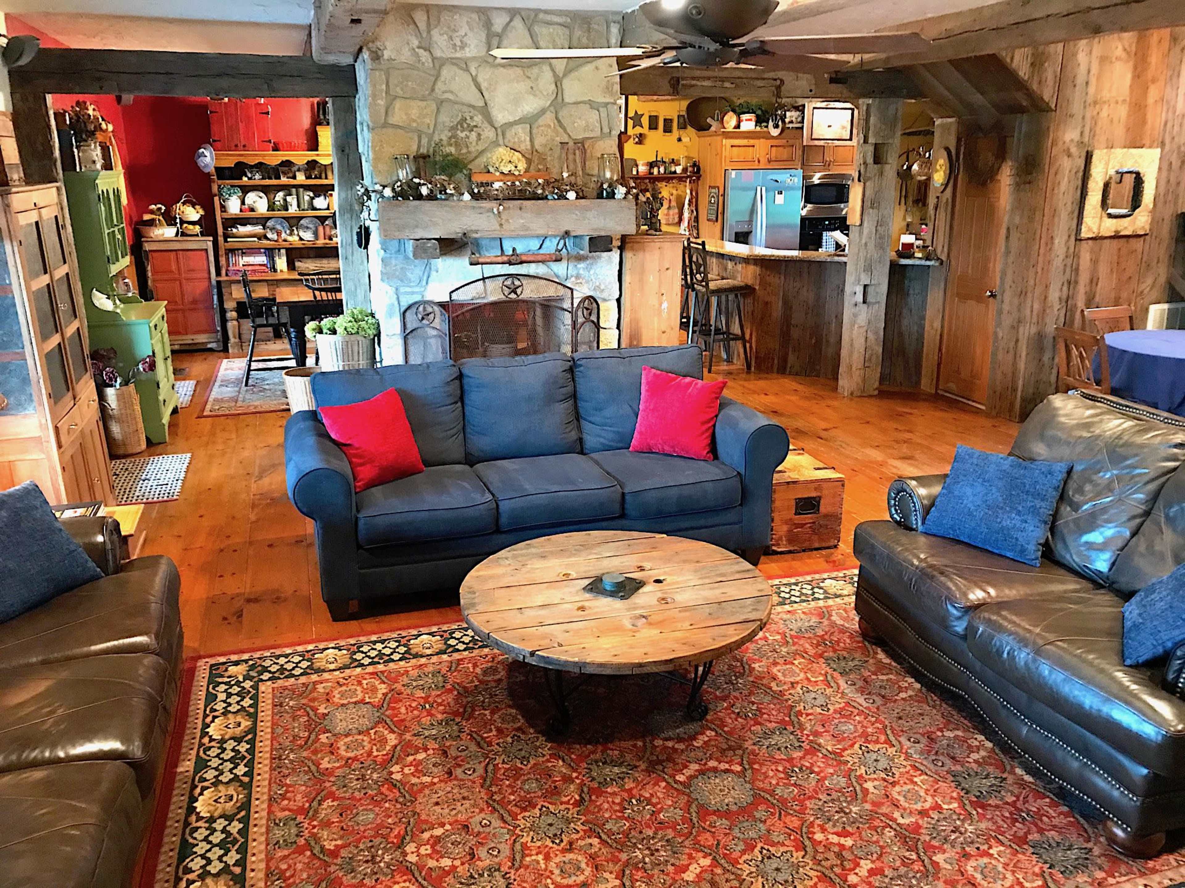 A cozy living room features a blue sofa with red cushions, a round wooden coffee table, and a stone fireplace, surrounded by wooden beams and rustic decor.