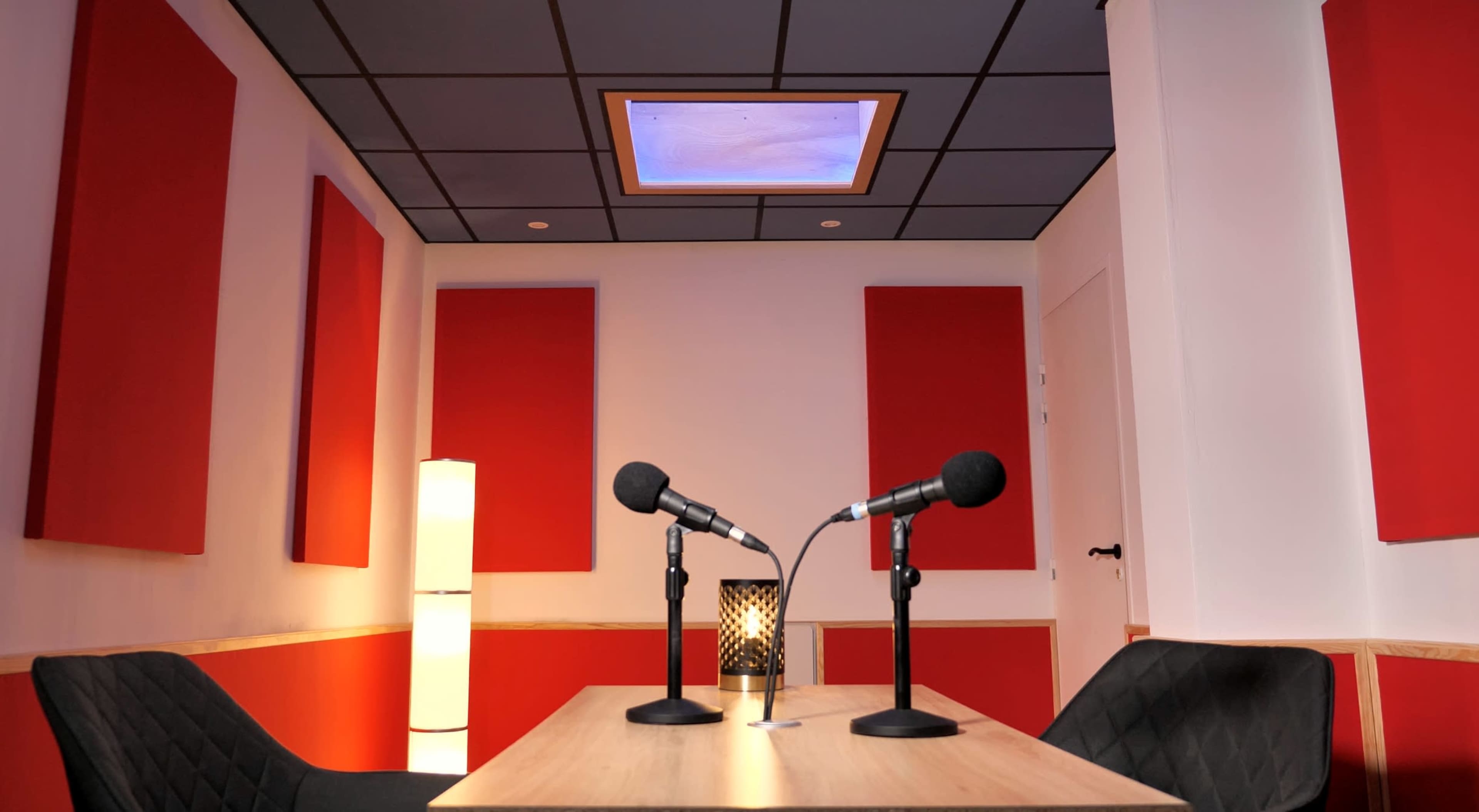 A soundproof recording studio featuring two microphones on a table, red acoustic panels on the walls, and a lamp in the corner.