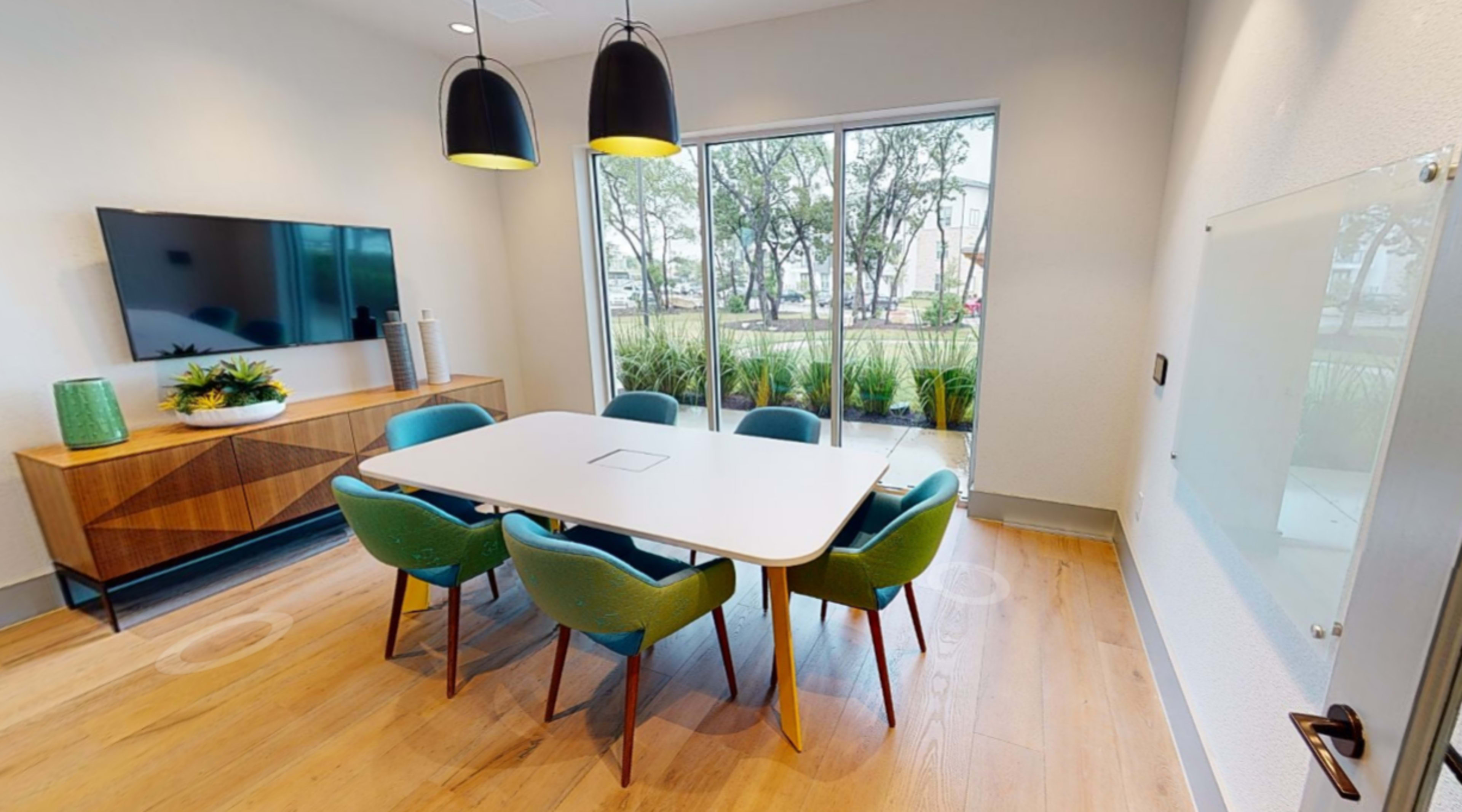 A modern meeting room features a rectangular table surrounded by colorful chairs, with a television mounted on the wall and large windows overlooking greenery outside.